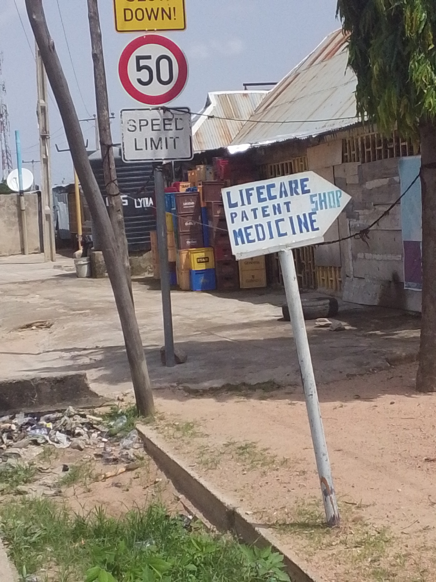 The image depicts a street scene in Jalingo, Nigeria, focusing on a directional sign. The sign, white with blue lettering, points to the right and reads "LIFECARE PATENT SHOP MEDICINE". Above the directional sign, there are two other signs: a red circular sign with "50" in white, indicating a speed limit, and a yellow rectangular sign with black text reading "SLOW DOWN!".
The background shows a row of buildings with corrugated iron roofs, suggesting a commercial or residential area. To the left of the signs, a large black tank with "S LYMA" printed on it is visible. In the foreground, the ground is paved with concrete, with patches of dirt and grass. A pile of garbage is visible in the lower left corner of the image. The scene appears to be outdoors, under daylight conditions.