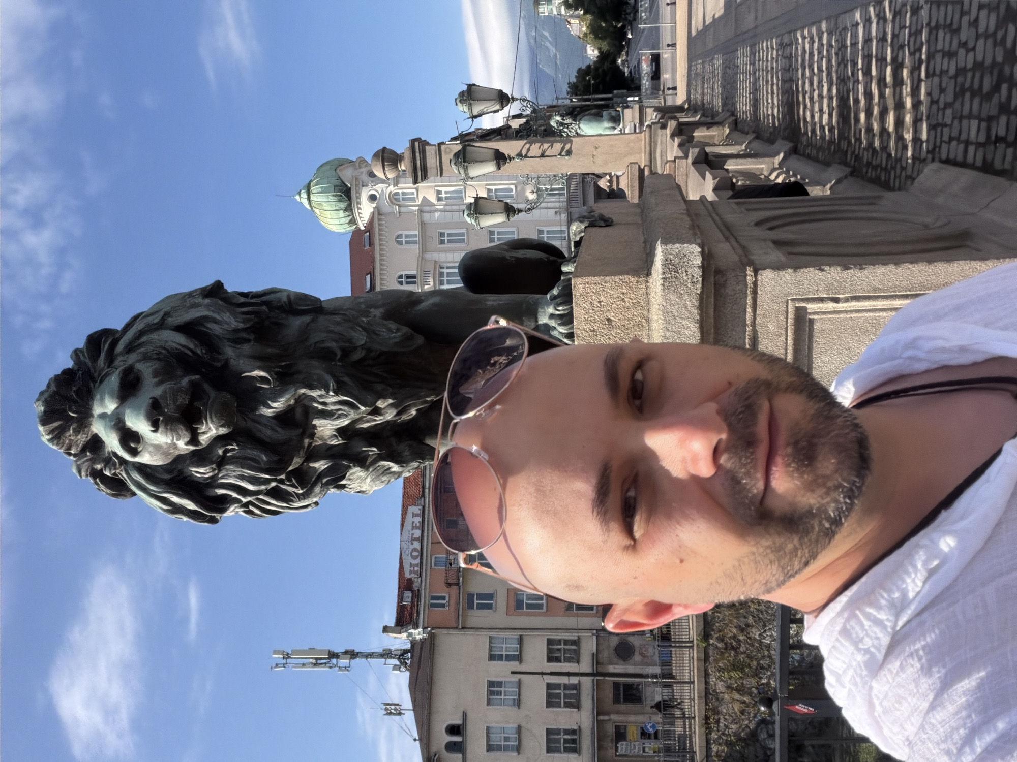 In Sofia, Bulgaria, a man with a bald head and sunglasses perched on his forehead takes a selfie on a bridge. Behind him, a large bronze lion statue stands guard, its mane detailed and imposing. The man is wearing a white hooded shirt. In the background, several buildings are visible, including one with a sign that reads "HOTEL". A green domed building is also present, and a street with cars and streetlights stretches into the distance. The sky is blue with some scattered clouds, and sunlight casts shadows on the cobblestone pathway of the bridge.