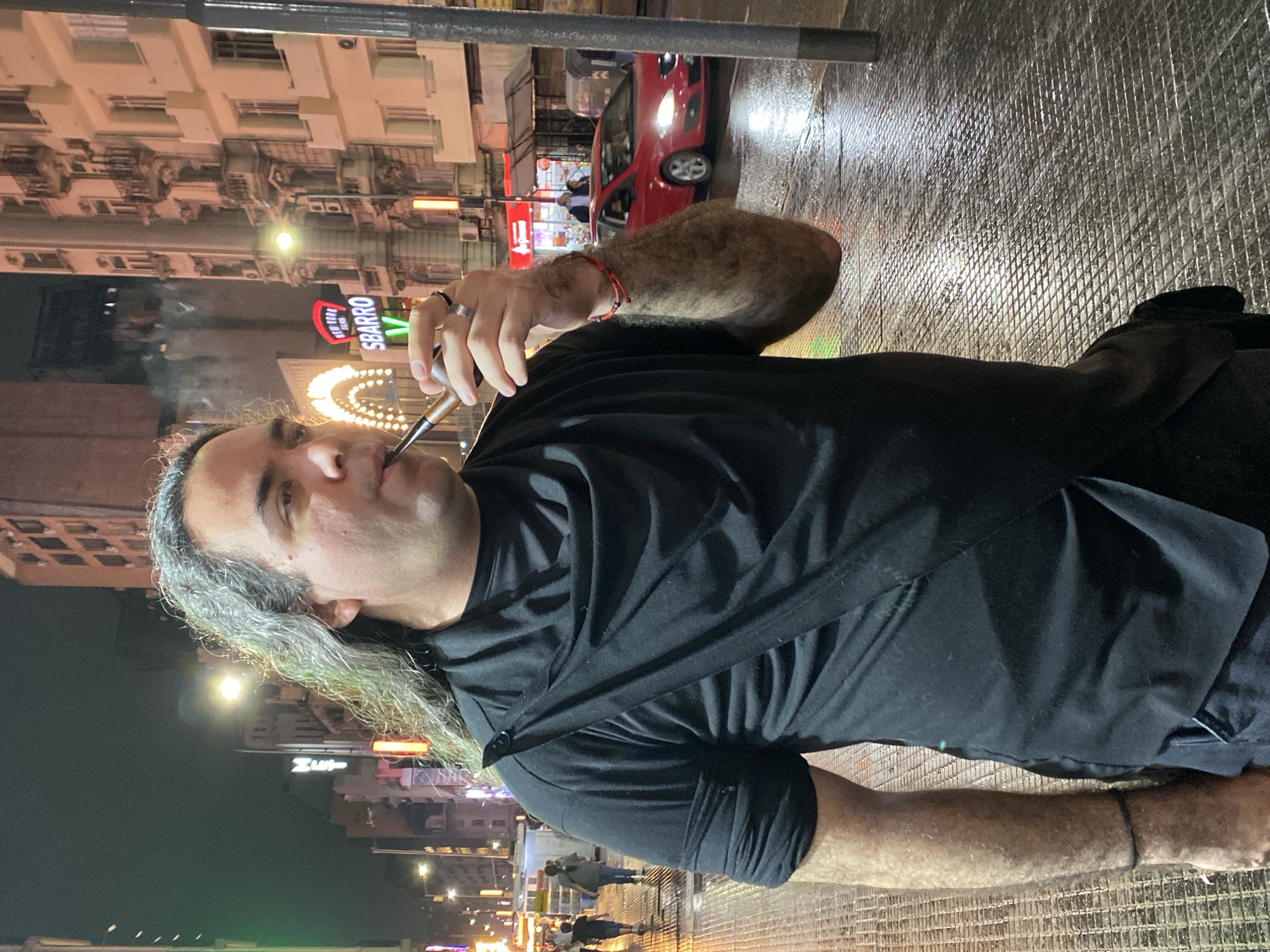 This nighttime image depicts a male individual smoking a pipe on a wet, tiled sidewalk in Buenos Aires, Argentina, near a Sbarro pizzeria.
The primary subject, a man with shoulder-length, wavy, grey-streaked dark hair, is seen in a right-facing profile, holding a dark-colored pipe with a lighter wood stem to his lips with his right hand. A plume of white smoke emanates from the pipe area. He wears a black, short-sleeved t-shirt featuring a draped, cowl-neck design with a diagonal fabric strap secured by a single button on his left shoulder. His right arm is visibly hairy, and he wears a silver ring on his right ring finger, two red string bracelets on his right wrist, and a black string bracelet on his left wrist.
The urban environment is illuminated by numerous commercial signs and streetlights, reflecting brightly off the wet, grey, grid-patterned pavement. A prominent "SBARRO" neon sign with "NEW YORK STYLE PIZZA" above it, accompanied by a curved array of yellow lights, is visible in the mid-ground. A vertical "MET" neon sign is also discernible on a building to the left. Multi-story buildings with architectural details, including ornate balconies on the right, line the street. Several blurred pedestrians are visible further down the sidewalk on the left, while a red four-door sedan drives on the street to the right.
