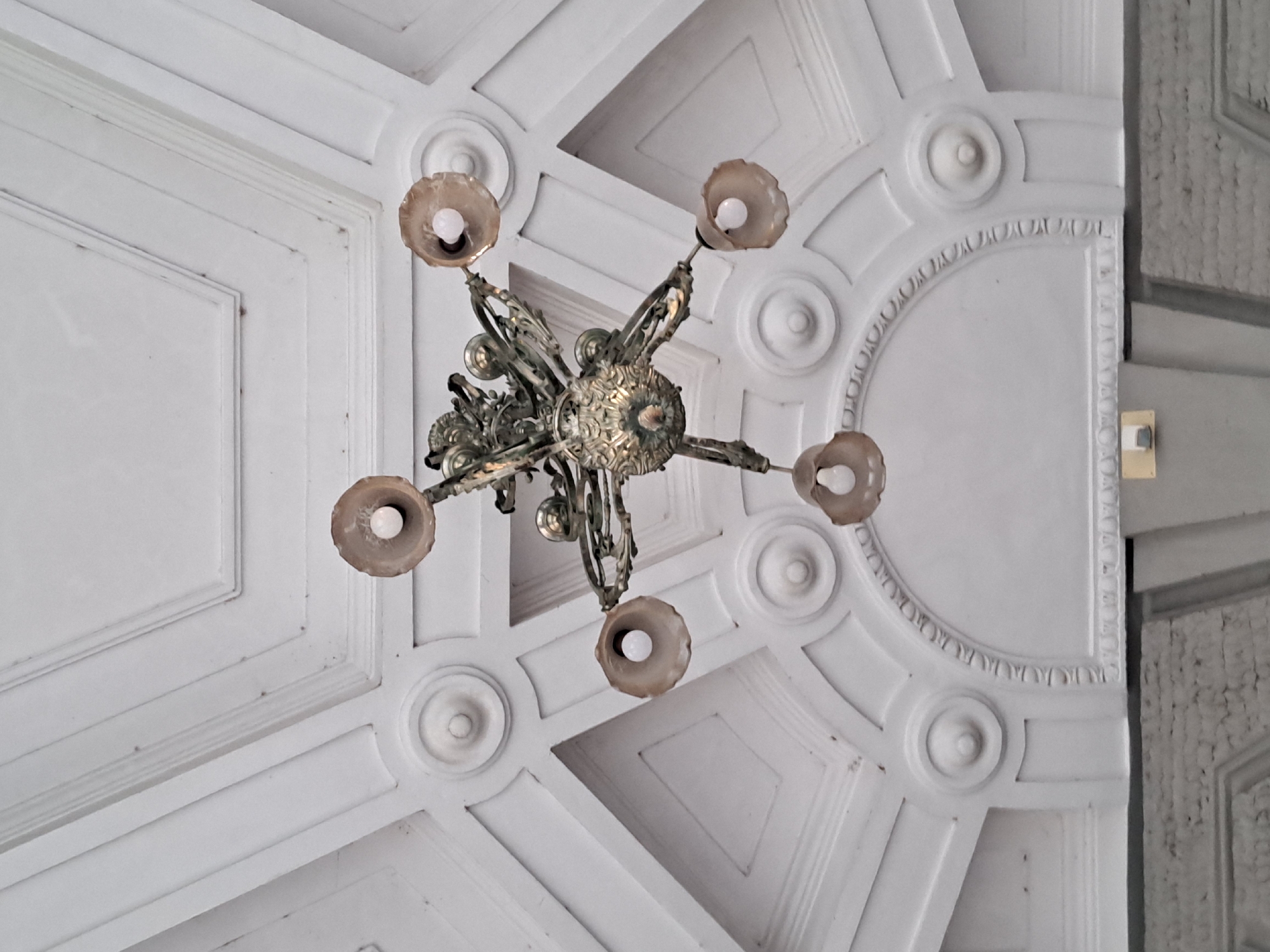 An ornate, antique chandelier hangs from a white, intricately molded ceiling in an interior setting. The chandelier is made of dark metal with five arms, each holding a small, decorative light fixture with a bulb. The ceiling features a geometric pattern of raised molding, including recessed squares, circular embellishments, and a large archway detail. The overall lighting is bright, illuminating the detailed architecture of the ceiling.
