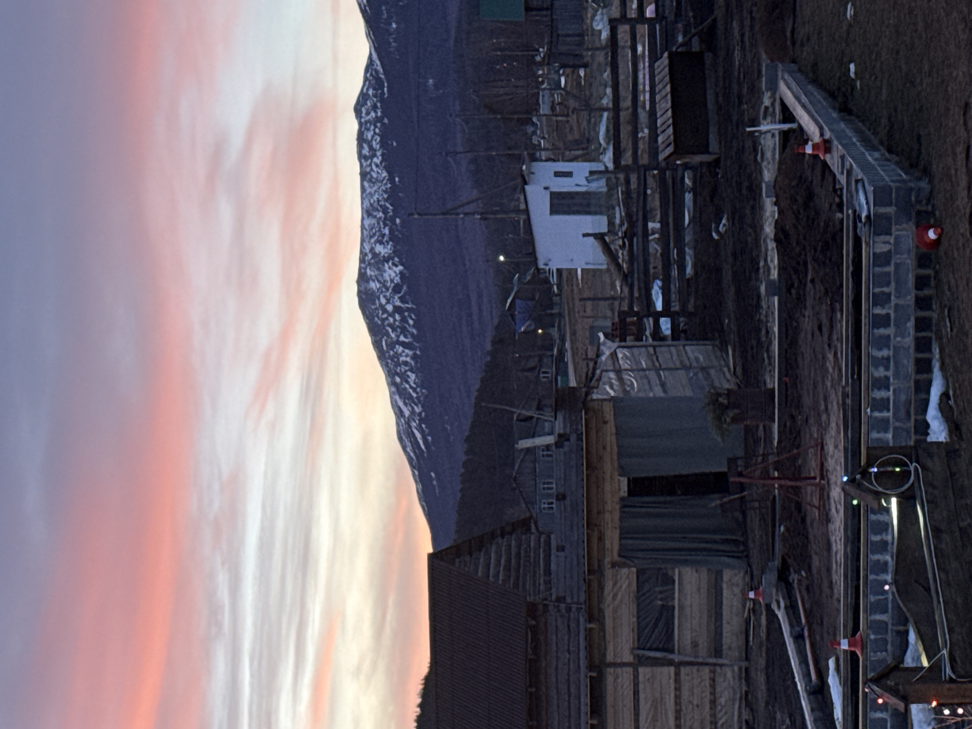 The image depicts a rural scene in the city of Tyulyuk, Russia, at twilight, characterized by a sky displaying hues of pink, orange, and purple transitioning into a lighter grey-blue, with scattered clouds. In the background, a snow-capped mountain range is visible under a darker, forested lower slope. The midground features a cluster of structures, including a prominent log cabin-style building with a dark, corrugated roof to the left. Further back, a small white utility building with a dark door stands amidst other wooden structures and wooden utility poles. The ground in the midground and foreground is bare earth with sparse patches of snow. In the foreground, a construction site is visible, featuring a rectangular foundation constructed from concrete blocks. Within and around this area, disturbed earth, several red and white traffic cones (some upright, some overturned), and construction debris are present. A dark wooden structure, possibly a planter or raised bed, adorned with string lights, is in the immediate foreground, alongside another small wooden decorative item also with string lights. A rusted barrel containing greenery is positioned near the construction zone. Wooden fencing delineates areas, and a dark, rectangular wooden box-like structure is behind one of these fences. No individuals or discernible animals are present in the scene.