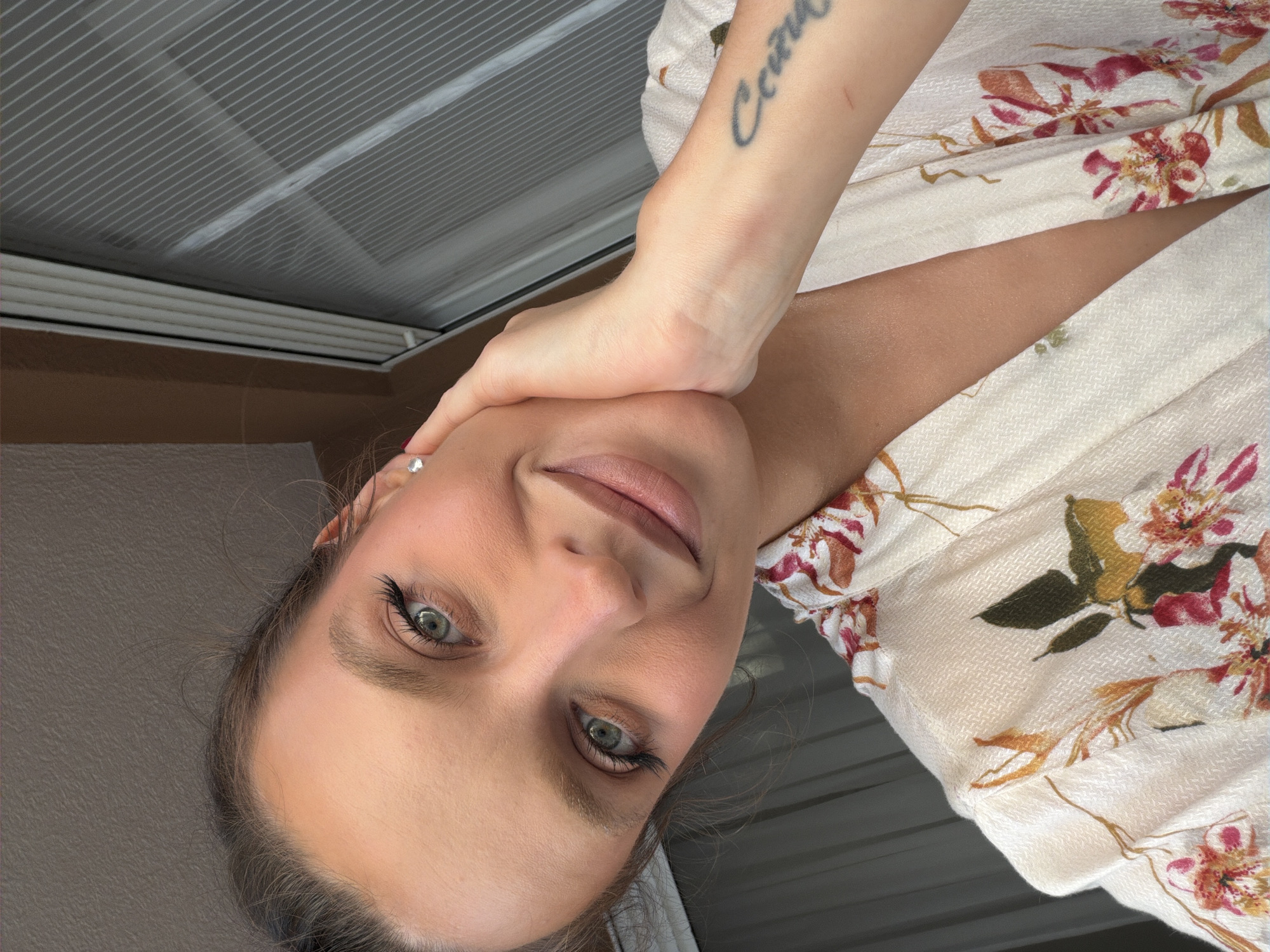 A woman with olive skin and green eyes poses for a selfie. She is wearing a light-colored, floral-patterned shirt and has a tattoo on her forearm. The background includes a window with blinds and a neutral-colored wall. The location cannot be definitively determined as Budva, Montenegro from the information provided.