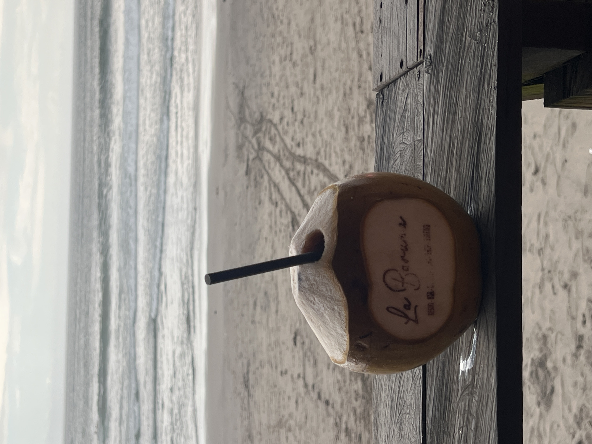 A refreshing coconut drink, complete with a black straw, rests on a textured, weathered wooden surface. The coconut husk is partially visible, and on its side, the stylized text "Le Barrus" is prominently displayed, with smaller, lighter text below it that reads "BALI SURFING" and a phone number (08570 8286 210).
The setting is a tranquil beach in North Kuta, Indonesia. In the blurred background, the calm ocean extends towards the horizon under a soft, possibly overcast sky, suggesting diffused daylight. Gentle waves lap at the sandy shore, which shows subtle patterns and indentations. The wooden surface appears to be a part of a deck, table, or railing, offering a vantage point over the beach. The overall scene conveys a sense of peaceful relaxation and tropical ambiance.