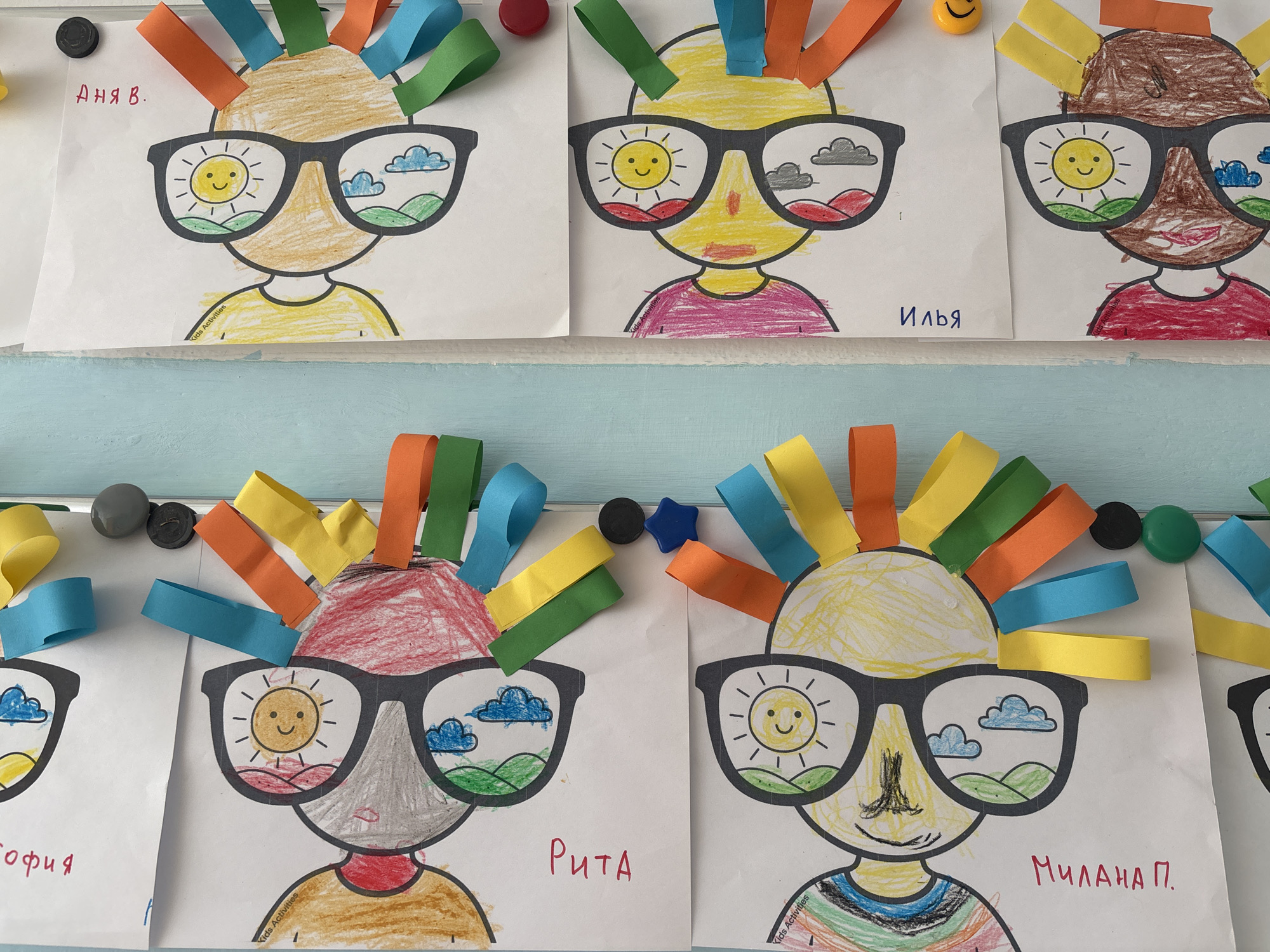The image captures a vibrant display of children's artwork, likely from a classroom or educational setting in Chaikovsky, Russia. The collection features several stylized portraits or faces, each characterized by large, dark sunglasses and a crown of colorful paper strips representing hair. These paper strips, curled and cut, come in shades of orange, yellow, green, and blue.
Each drawing depicts a head and shoulders, with varied skin tones and different colored shirts, indicating individual creative input from the young artists. A prominent feature is the reflections within the sunglasses lenses, which showcase diverse weather and landscape elements. Some lenses display a cheerful yellow sun, while others show blue or grey clouds, and some include green rolling hills. Often, one lens presents a sunny scene while the other shows clouds, suggesting varied perspectives or moods. Small text, "Kids Activities," is visible on several drawings, implying they are part of a structured project.
The artwork is fastened to a white surface, possibly a whiteboard or wall, using an assortment of magnets. These magnets vary in color and shape, including black, grey, red, a yellow smiley face, a green button, and a blue star. A light blue border or shelf runs along the bottom edge of the display.
Visible names written in Cyrillic script identify some of the artists: "АНЯ В." (Anya V.), "ИЛЬЯ" (Ilya), "СОФИЯ" (Sofia), "РИТА" (Rita), and "МИЛАНА П." (Milana P.). The display radiates a cheerful and creative atmosphere, showcasing the children's imaginative interpretations.