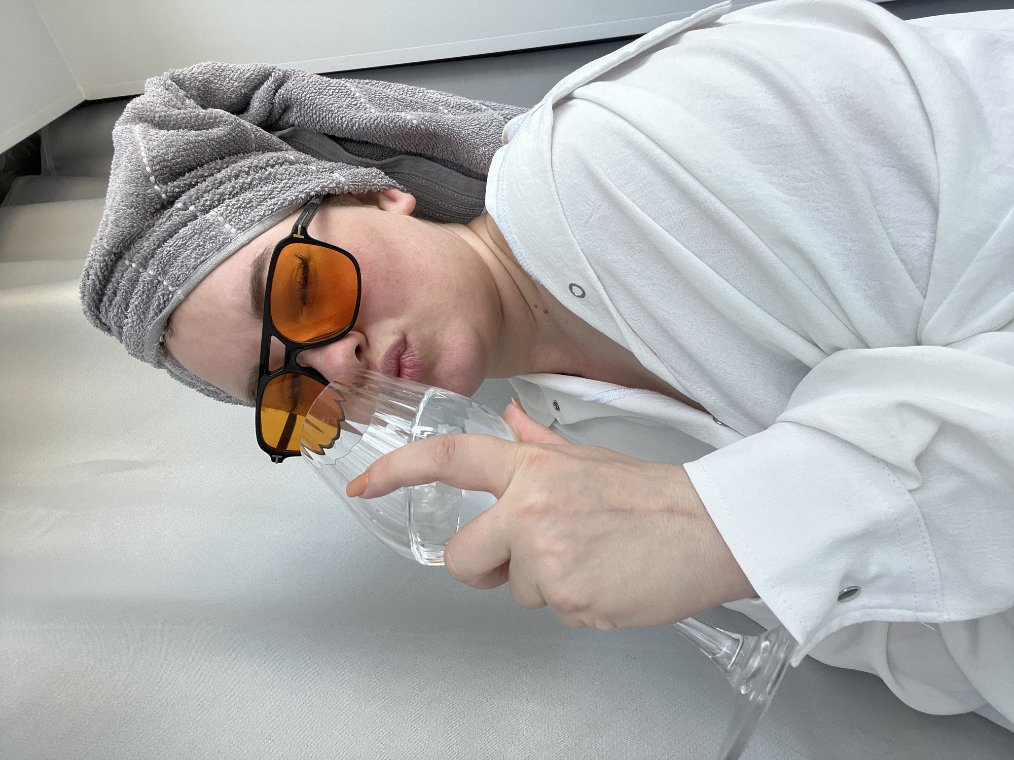 A person wearing orange-tinted glasses and a gray towel turban is lying down. They are wearing a white, button-down shirt and holding a clear glass. The setting appears to be indoors, possibly in Chaikovsky, Russia, but specific city details are not evident from the image.