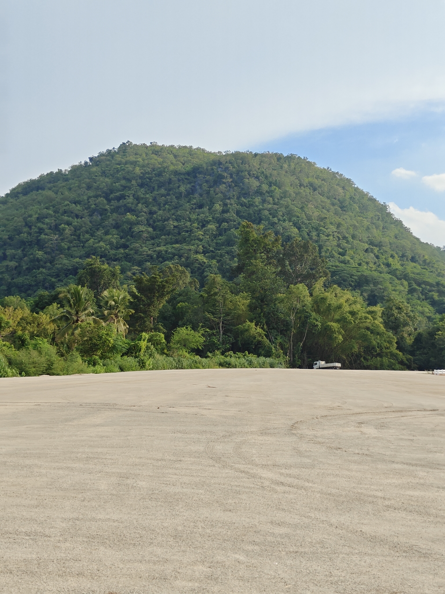 The image depicts a wide, flat expanse of light-colored gravel in the foreground, with tire tracks visible across its surface. In the middle ground, a dense line of lush green trees, including palm trees and bamboo, forms a visual barrier. Behind the trees, a large, verdant mountain dominates the background, its slopes covered in thick vegetation. A small white truck is parked near the edge of the tree line. The sky above is a pale blue with a few wispy white clouds. The scene appears to be an outdoor area, likely a clearing or a parking lot adjacent to a natural landscape, situated in the vicinity of Mu Si, Thailand, a region known for its scenic natural beauty and mountainous terrain.