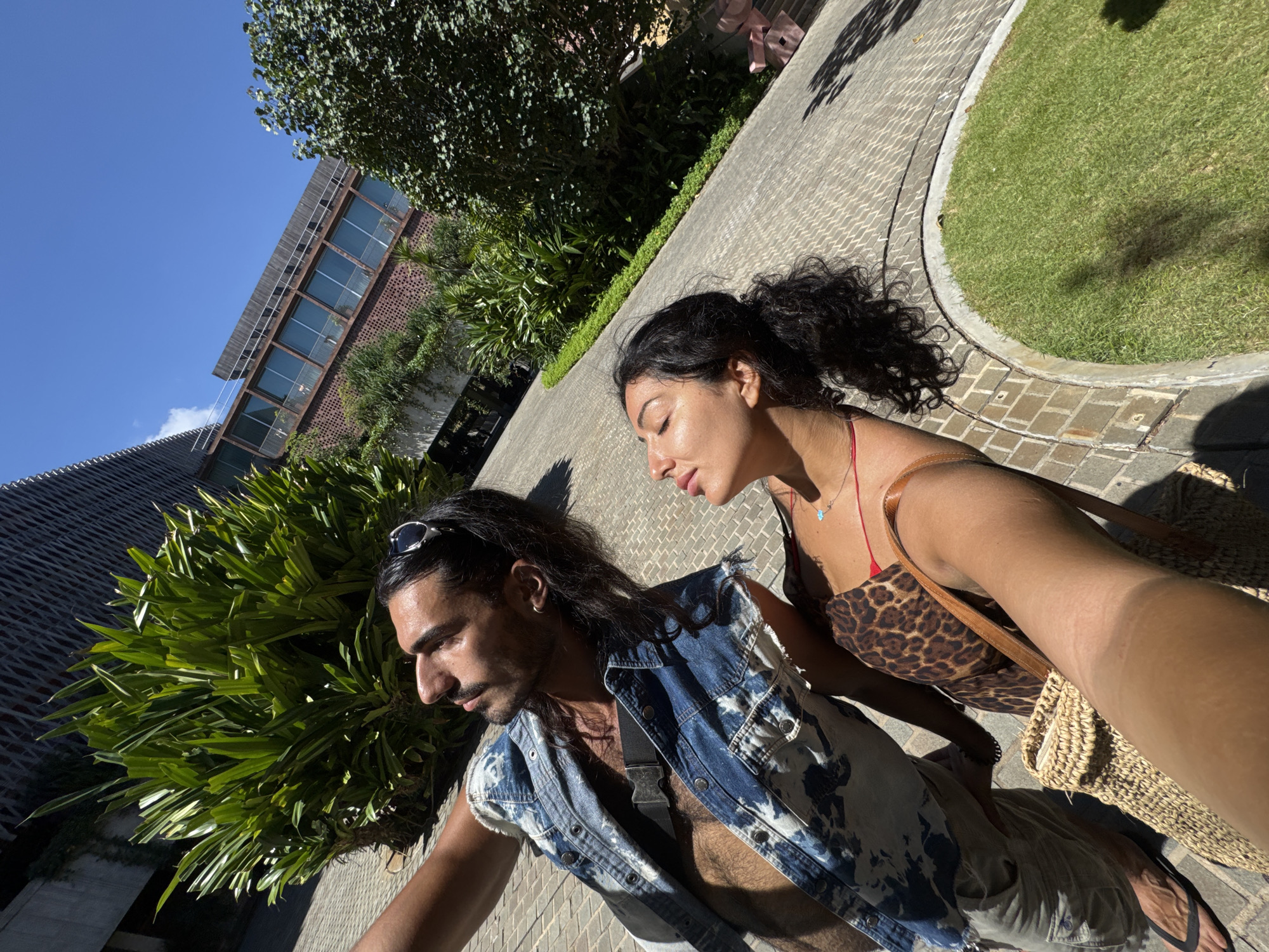 The photograph, taken at an oblique angle, depicts a man and a woman in an outdoor setting. The man, on the left, has dark hair pulled back from his face, sunglasses perched on his head, and a mustache and beard. He is wearing a blue and white sleeveless denim jacket over what appears to be a dark shirt. His bare chest is partially visible. The woman, positioned to the right, has dark hair pulled up in a ponytail. She is wearing a leopard-print dress with a red strap visible, and a tan bag slung over her shoulder. The background includes a textured brick building on the left, lush green foliage in the center, and a paved pathway with a patch of grass on the right. The sky is bright and clear. The photograph was taken in North Kuta, Indonesia.