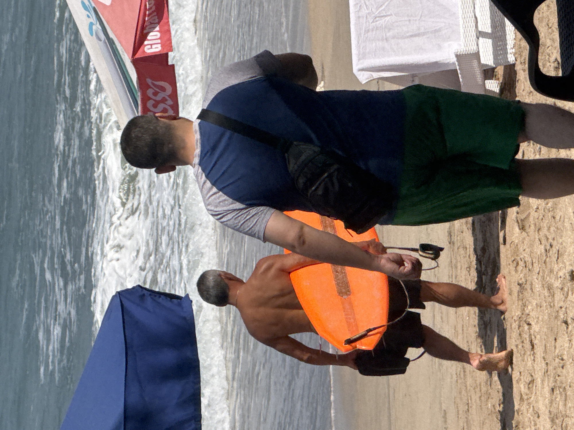 The image depicts a beach scene in Kuta, Indonesia, with two men on the sand. The man on the left, facing away from the camera, has short gray hair and is carrying an orange surfboard. He is wearing dark shorts and appears to be walking towards the ocean. The man on the right, also facing away, is wearing a blue and gray t-shirt and green shorts. He has a black crossbody bag slung over his shoulder. In the background, the ocean waves are visible, and a red and white striped umbrella is set up on the beach. Further in the background, another umbrella, this one blue, provides shade. There are lounge chairs in the foreground on the right side of the image.