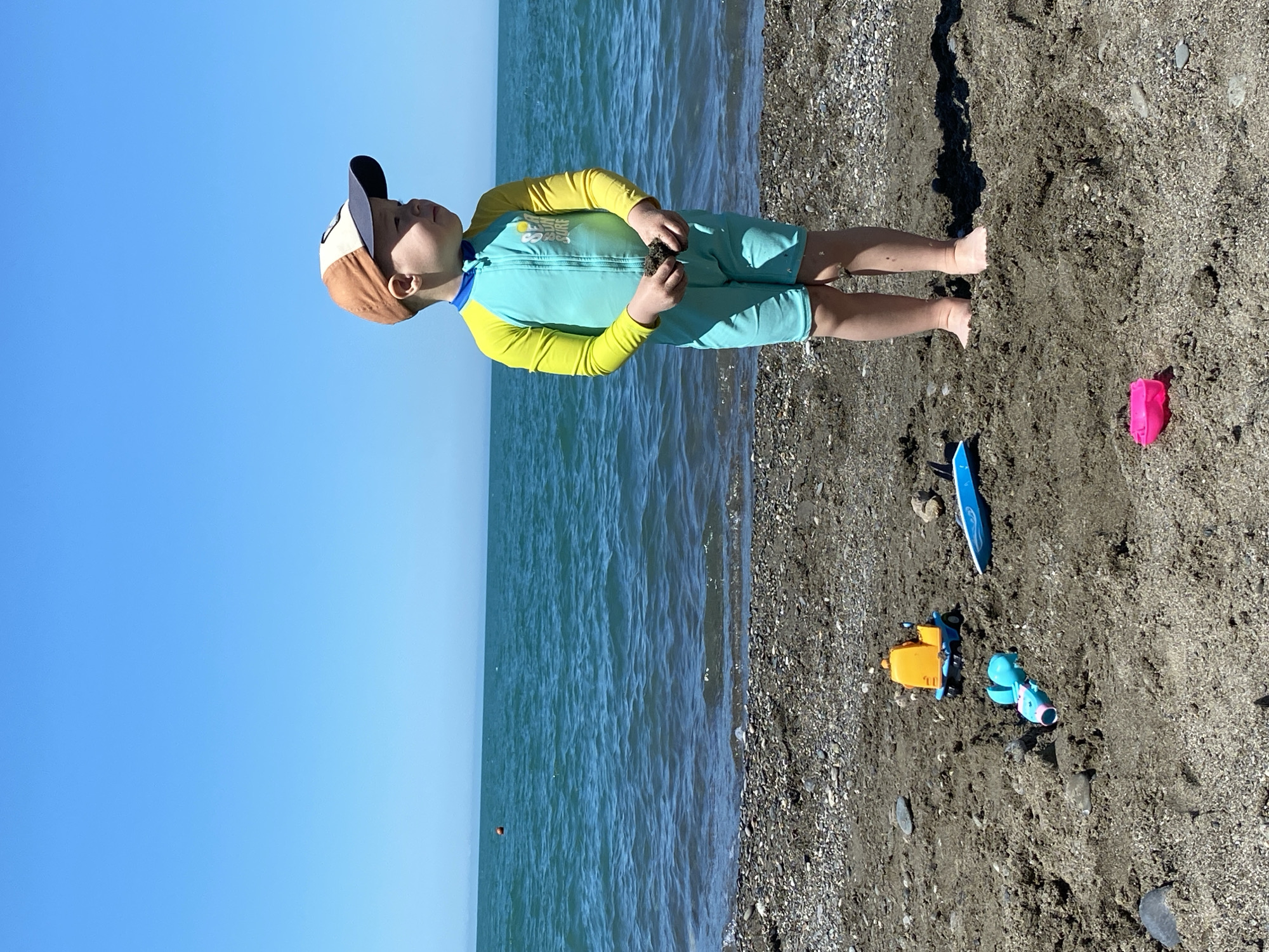 This forensic description details a bright, sunny scene at a pebbly and sandy beach in Sochi, Russia. A young child, appearing to be a toddler, stands barefoot on the dark sand, facing right in profile. The child wears a long-sleeved, short-legged turquoise and yellow rash guard with "SEA SUN SURF" printed on the chest, and a brown and navy blue baseball cap. Both hands are clasped, holding a small lump of dark sand. Various beach toys are scattered in the foreground: a yellow and blue toy truck, a light blue and pink toy shovel, a light blue toy surfboard, and a pink toy bucket. Behind the child, the calm, turquoise-blue sea extends to a distant, hazy horizon where a faint landmass is visible. A single orange buoy floats in the mid-left area of the water. The sky above is clear and brilliant blue, indicative of a bright, warm day.