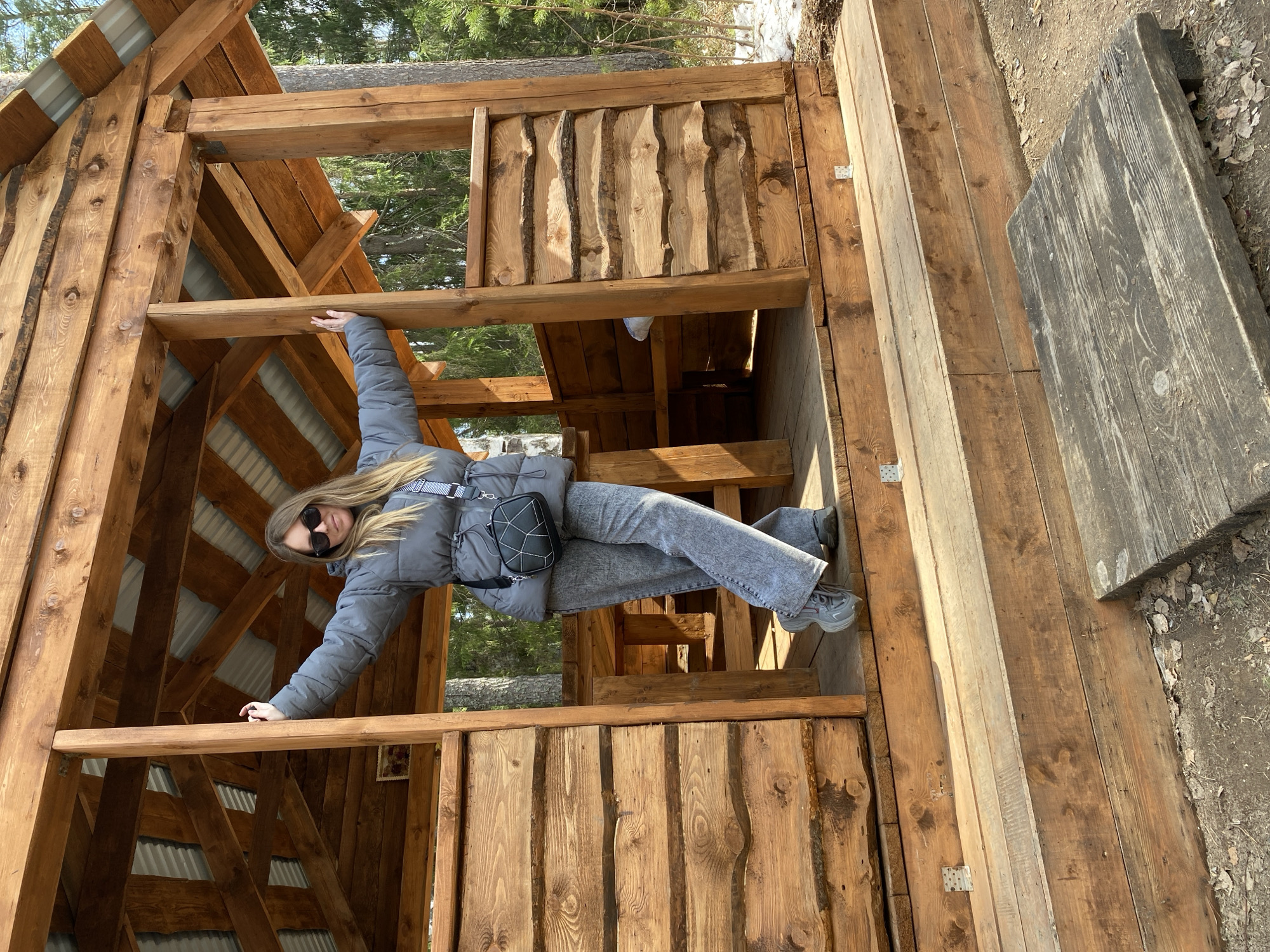 A young woman with long blonde hair, wearing a grey puffer jacket, grey jeans, grey sneakers, and black sunglasses, is posing inside a rustic wooden structure. She has a black crossbody bag with a white geometric pattern slung over her shoulder. Her arms are outstretched, with one hand resting on a horizontal wooden beam at shoulder height and the other on a lower beam. She looks directly at the camera with a slight smile.
The structure is made of rough-hewn wooden planks and beams, giving it a natural, unrefined look. Corrugated metal roofing is visible through the gaps in the wooden framework, suggesting either an unfinished building or an open-air shelter. The setting appears to be outdoors in a forested area, with green pine trees visible in the background. Patches of snow are visible on the ground outside the structure, indicating a cold or early spring season. The bright sunlight suggests it is daytime. The ground immediately outside the structure is covered with dirt, dry leaves, and some snow patches. No visible text is present in the image.
The location is Noviy, Russia.
