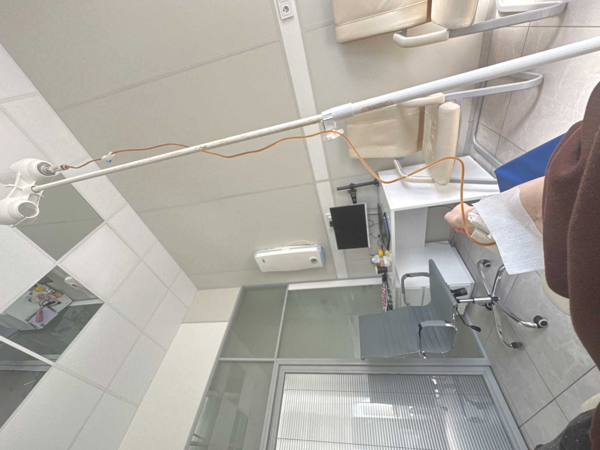 The image shows a medical setting, likely a clinic or hospital room. The perspective is from the patient's point of view, with an IV drip visible, inserted into their arm. The room contains medical equipment, chairs, and a desk with a computer screen. A window with blinds is visible. There are no details that indicate this is located in Moscow, Russia. The location cannot be confirmed based on this image.