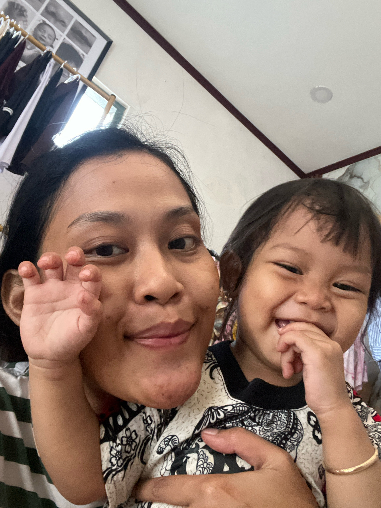 In an indoor setting, a woman and a young child are positioned in a close-up, selfie-style shot. The woman, on the left, has dark hair and is looking towards the camera, holding up her right hand. The child, on the right, is smiling broadly and has their hand near their mouth. They are both wearing patterned clothing. The background shows parts of a room, including a clothes rack with garments and a framed picture on the wall. The lighting suggests it is daytime. The context of Kecamatan Pati, Indonesia is not discernible from the visual information provided in the image.