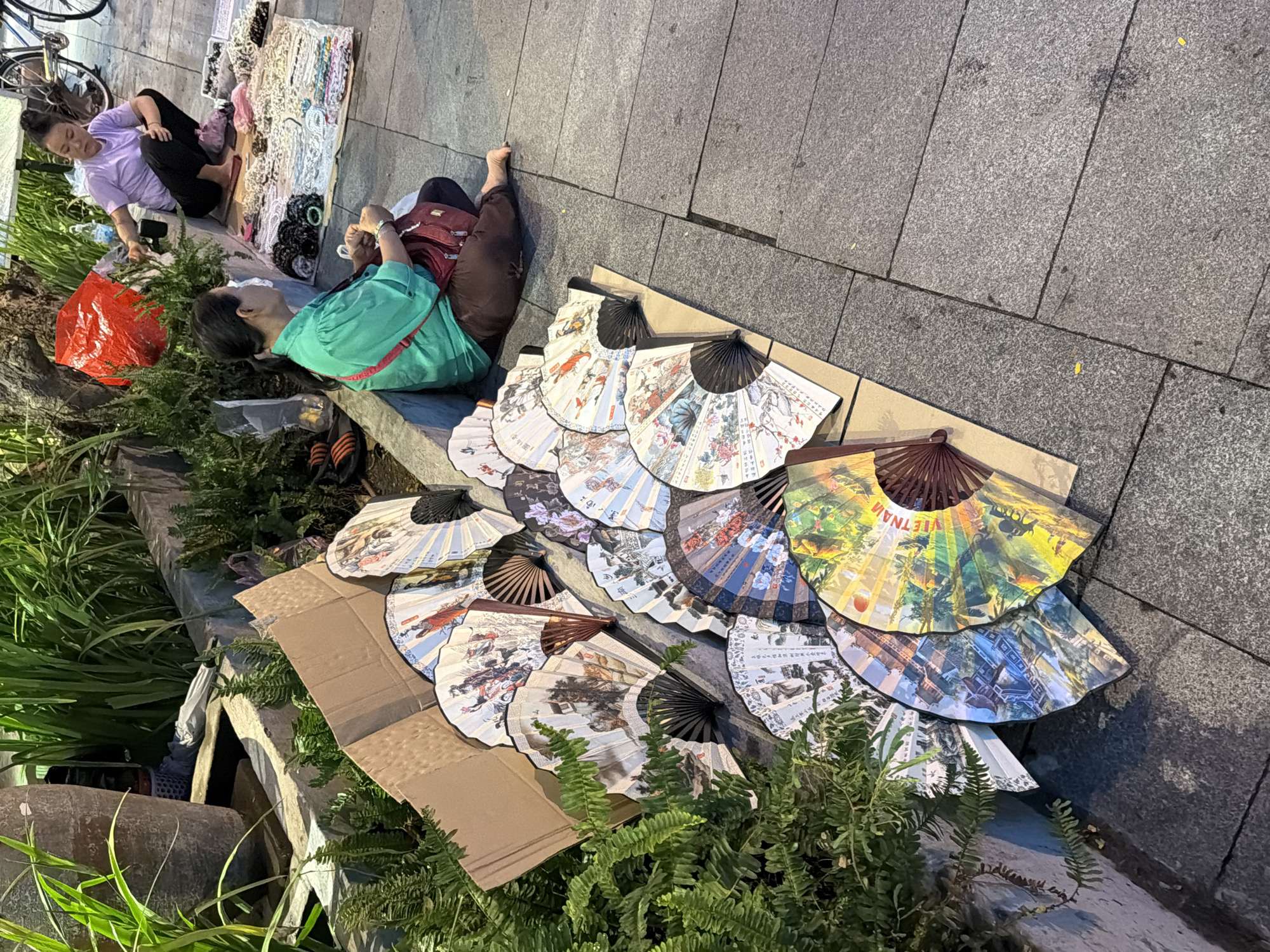 The image depicts an outdoor market scene in Nha Trang, Vietnam, where two women are selling goods. In the foreground, a collection of decorative fans, many with intricate designs of people and landscapes, are spread out on cardboard and the pavement. To the right, a woman in a green shirt is seated on the ground, attending to her merchandise. Further back, another woman in a light purple shirt is crouching, also engaged in selling. A variety of accessories, possibly jewelry or trinkets, are laid out on a mat in front of her. The background reveals lush greenery, a large concrete planter, and a bicycle, suggesting a public urban setting. The ground is paved with grey stone tiles.