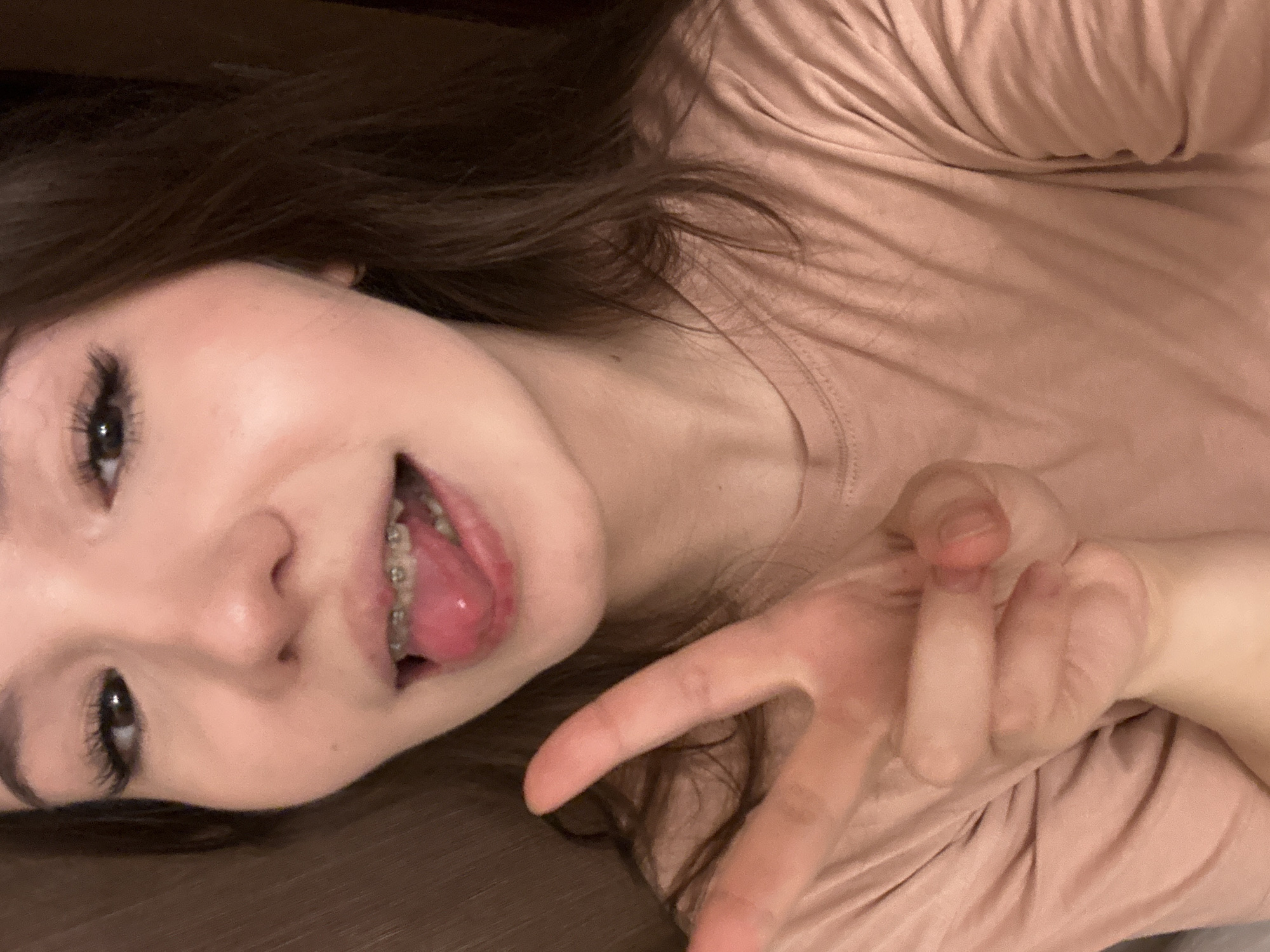 A young female subject with long, dark brown hair is depicted in a playful, close-up portrait, seemingly taken from above as she reclines or lies down. She wears a light peach or blush-colored t-shirt. Her head is tilted, and she looks directly at the camera with a casual, possibly mischievous expression. Her mouth is open, revealing her tongue extended slightly and metal braces on her upper teeth. Her right hand is raised in the foreground, making a peace sign (V-sign) with her index and middle fingers.
The setting appears to be indoors, likely a relaxed home environment such as a bed or sofa, given the crumpled fabric visible on the right side and the soft, warm lighting. The background is dark and out of focus, providing little specific detail about the room. The overall mood is candid and lighthearted. There is no visible text in the image. This scene is set in Naberezhnye Chelny, Russia.