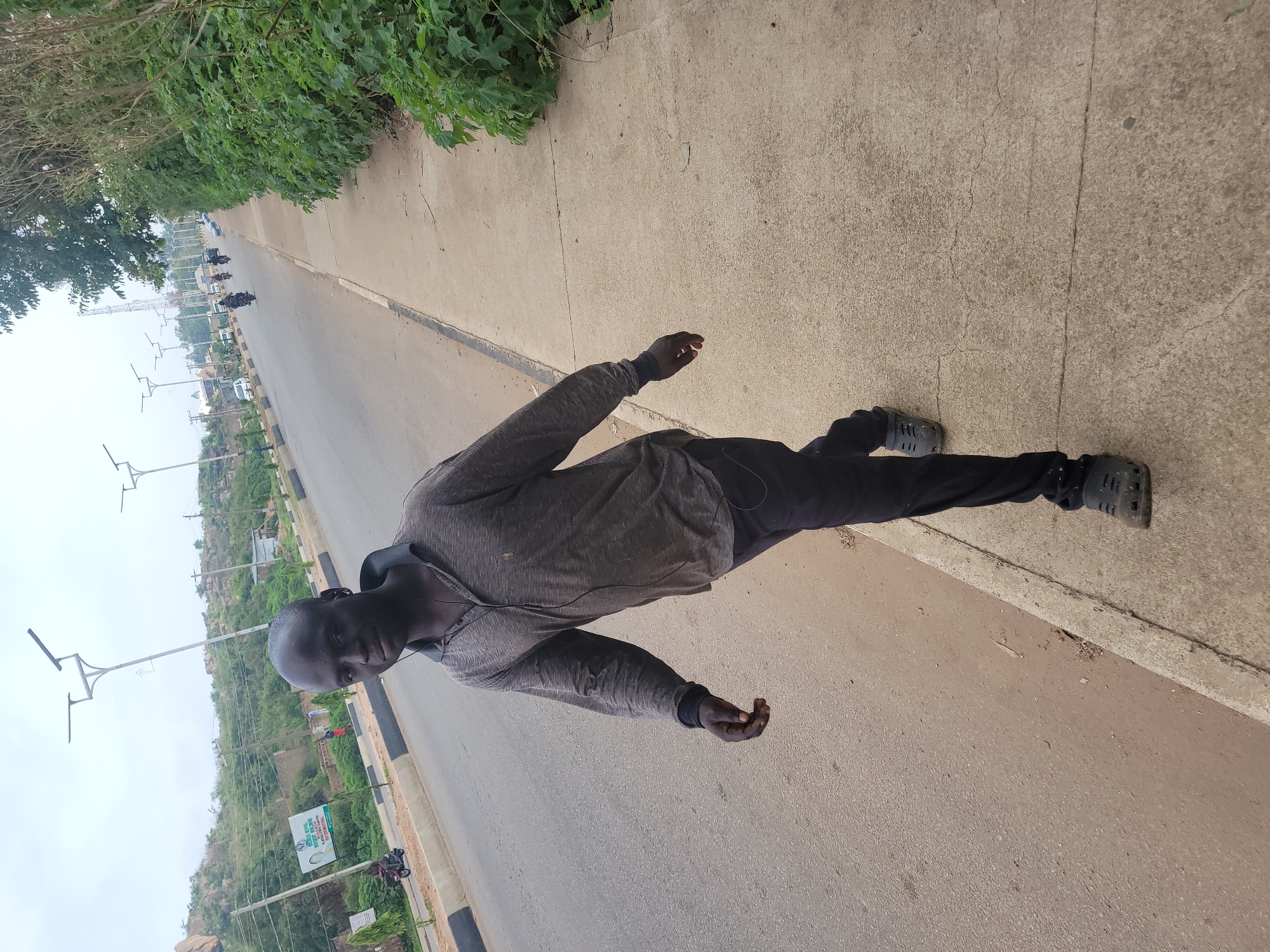 The image shows a man walking on a concrete sidewalk next to a paved road. He is wearing a long-sleeved gray shirt and black pants, and dark-colored clogs. The man has short dark hair and is looking towards the camera. In the background, there is a road with several motorcycles and cars, a billboard with text in English and possibly another language, trees, and rocky hills. The sky is overcast. The scene is consistent with a street view in Gura Top, Nigeria, given the architectural style of the lampposts and the general environment.