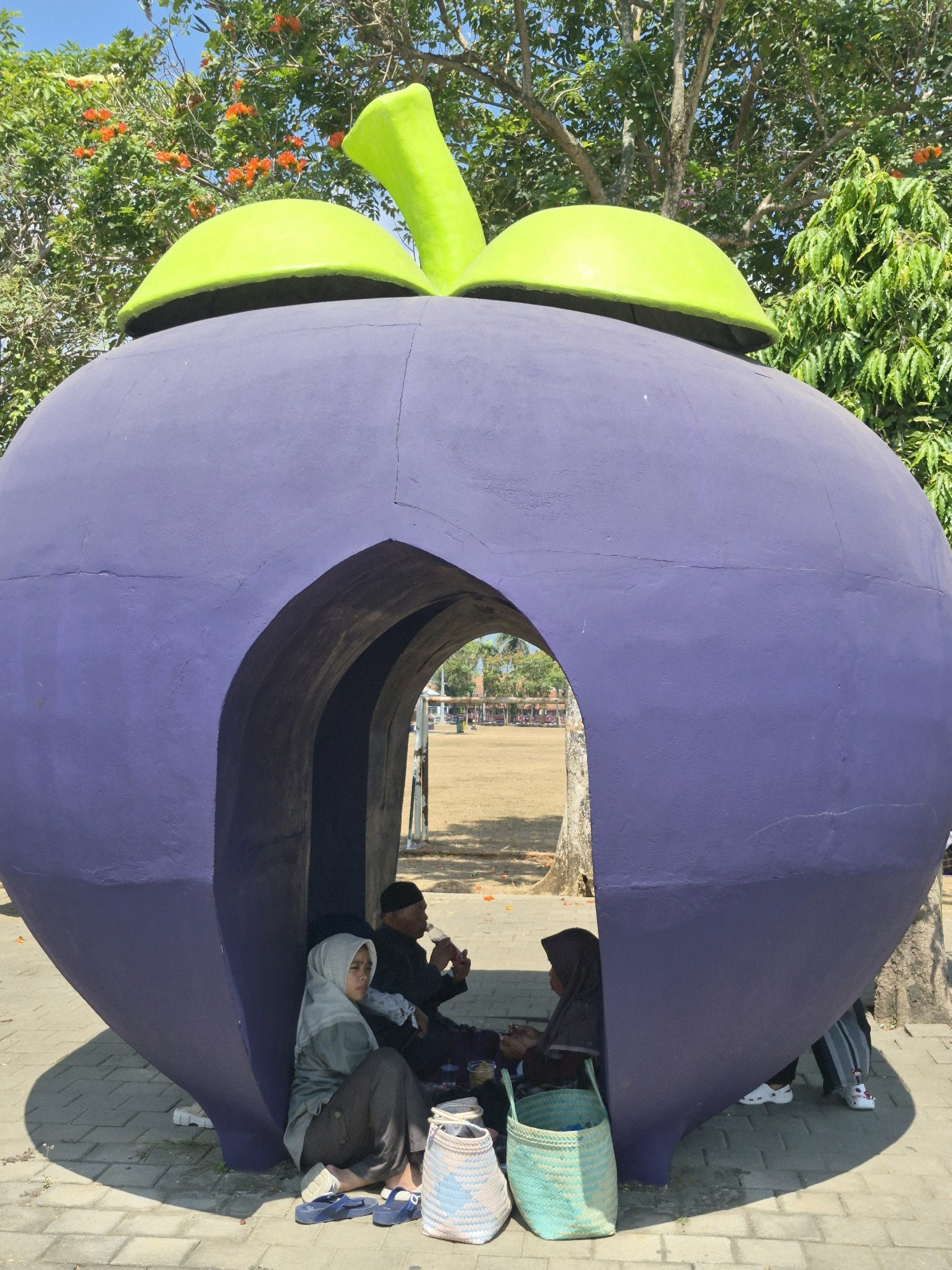 The image depicts a large, purple, mangosteen-shaped structure with a green stem and cap, serving as an entrance for people to sit inside. Three individuals are visible within the opening of the structure. On the left, a person wearing a light gray hijab and dark pants is seated on the ground, leaning against the purple structure. To their right, a person in a black cap and dark clothing is seated, holding a bottle and drinking. Further to the right, another person wearing a dark hijab and dark clothing is seated. Two woven baskets, one light blue and white and another light green, are placed on the ground in front of the seated individuals. The structure is situated outdoors on a paved walkway, with trees and foliage visible in the background. Sunlight casts shadows on the walkway, indicating it is daytime. The overall scene appears to be a public space, possibly a park or recreational area in Purworejo, Indonesia, given the context of the question.