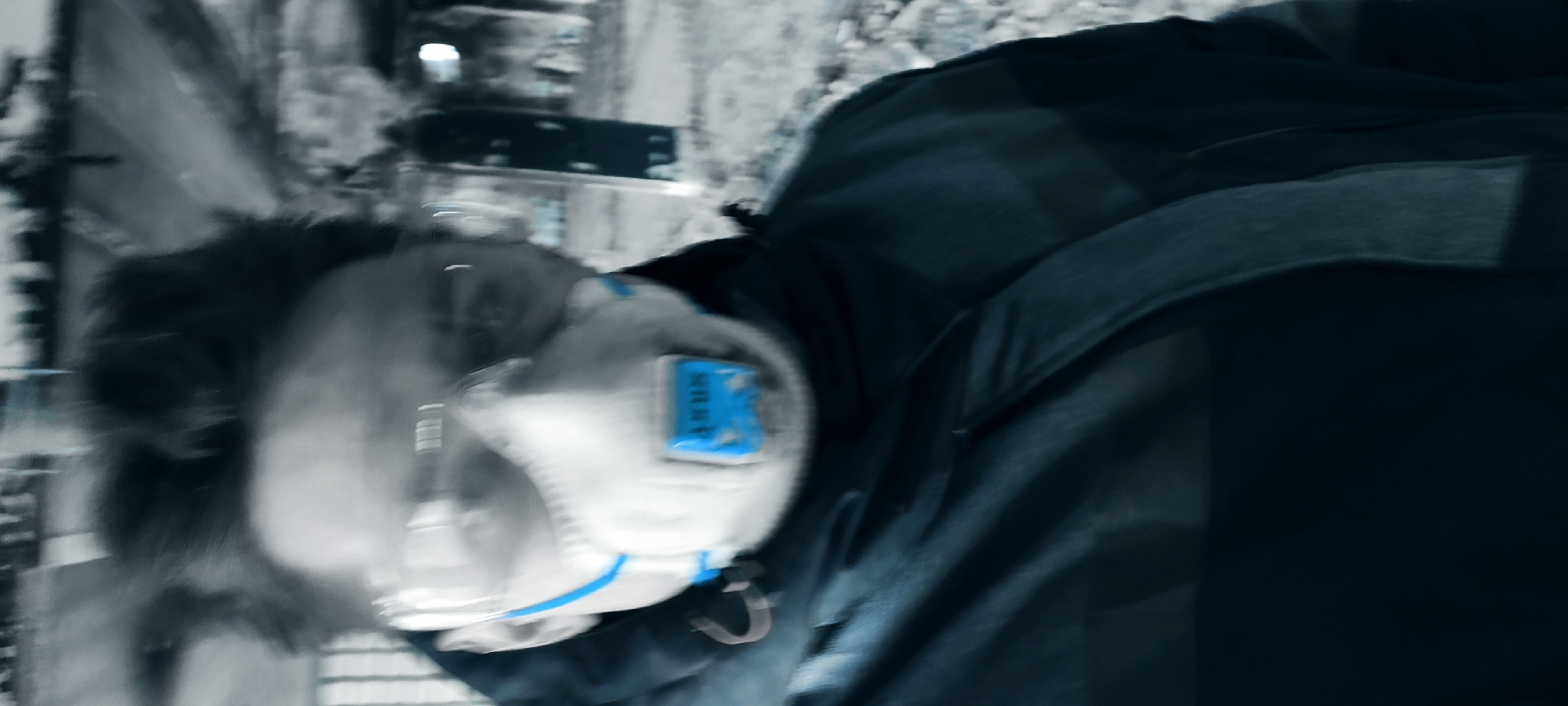 The image captures a person from a slightly tilted, close-up perspective, suggesting motion or an action shot. The individual, likely male with dark, slightly disheveled hair, is wearing a white protective respirator mask. This mask features a prominent blue rectangular valve or filter cover on the front, with faint, blurry white text that appears to read "NRR". A thin blue stripe runs along the lower edge of the mask. Over the mask, the person wears a pair of clear glasses, possibly safety glasses. They are dressed in a dark, potentially deep blue or black, jacket or coat, which occupies a significant portion of the frame.
The background is heavily motion-blurred and desaturated, rendered in shades of gray and muted blue, providing minimal detail. It vaguely suggests an outdoor urban environment with indistinct buildings or structures and possibly a street. The overall aesthetic of the image is monochromatic with a cool blue tint, making the blue elements of the mask and the dark clothing stand out. The significant blur throughout the image, particularly in the background and around the person's head, indicates rapid movement or a low-light condition. The time of day and specific weather conditions are not discernible. No clear location cues are visible within the image itself.