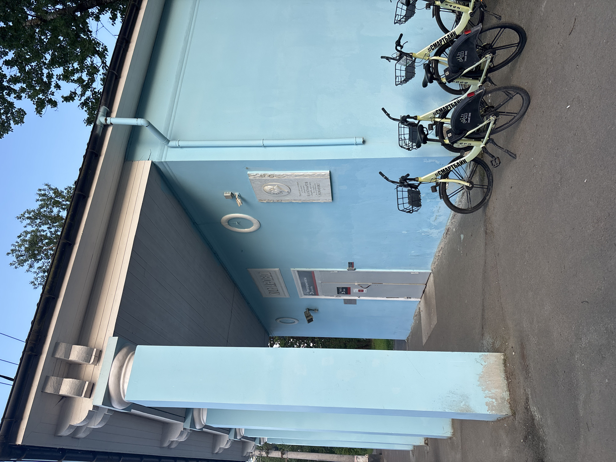 The image captures the exterior of a light blue building, likely a suburban ticket office or station, in Komarovo, Russia. The building features classical architectural elements, including prominent pillars and a decorative cornice along the roofline. A white drainpipe system runs along the upper part of the wall.
Parked against the building are three cream-colored rental bicycles, each equipped with a black front basket and mudguards. The bicycles are branded with the text "#СМАРТБАЙК" and "SMART BIKE", and some bear numbers like "1462". The bikes suggest a public bike-sharing or rental service is available at this location.
On the building's wall, a white commemorative plaque is visible, featuring a portrait and text that includes "В.Л. КОМАРОВ" and "КОМАРОВО". Below this, a white circular light fixture and a security camera are mounted. Further down, a light-colored door is visible, above which the text "Пригородные кассы" (Suburban ticket offices) is displayed, with some less clear text below it that OCR identifies as "T STARRE". Faintly visible on the underside of the awning is the word "КОМАРОВО".
The setting appears to be daytime with a clear, bright sky visible above a tree with green foliage on the left. The ground is paved with asphalt. No people are present in the image.