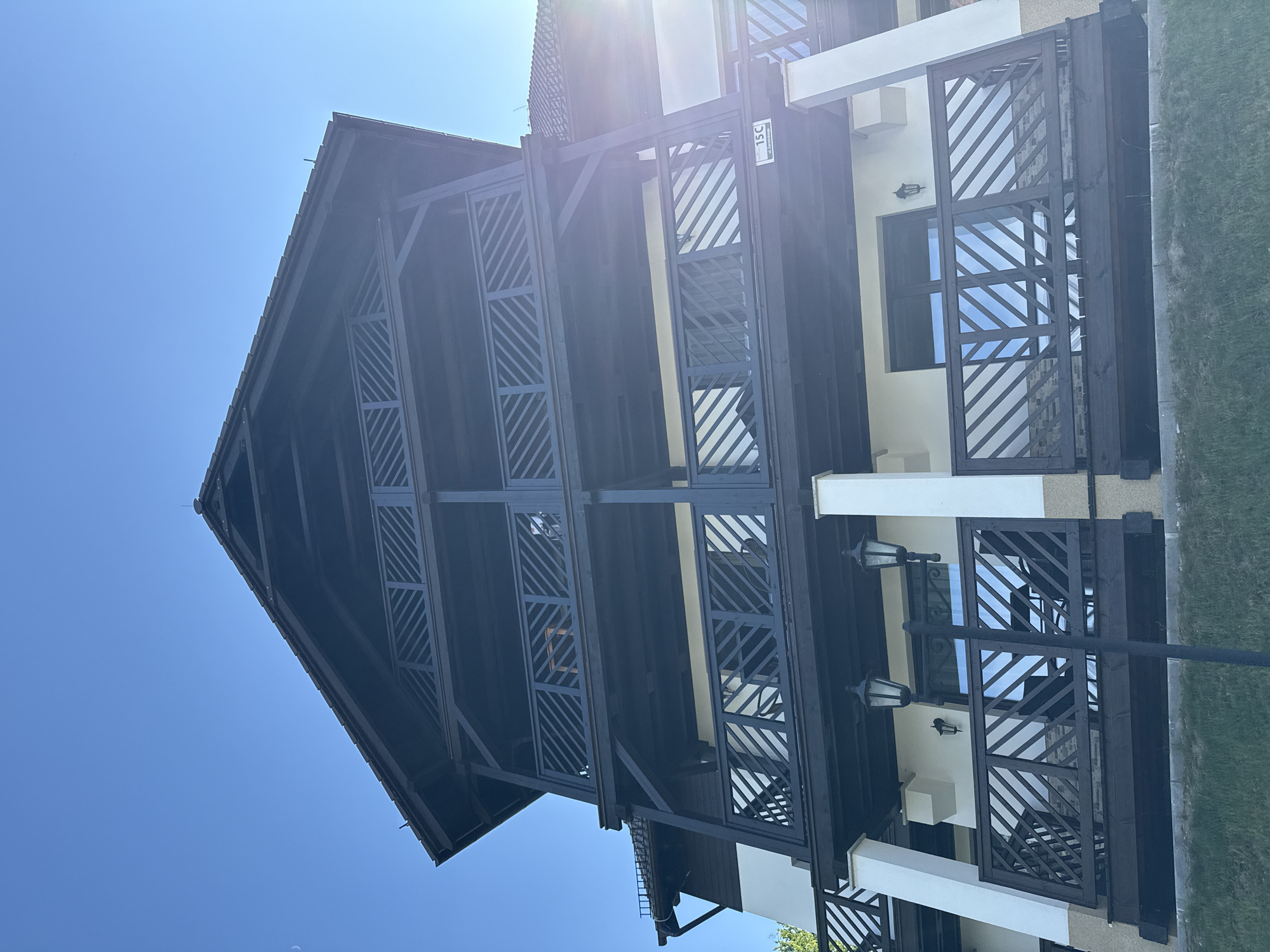 The image shows a multi-story building with dark wooden balconies and railings, set against a clear blue sky. The architecture appears to be in a chalet or lodge style. One balcony has a visible sign with "15C" and "ul. Żeromskiego" written on it, indicating an address on Żeromskiego Street in Szklarska Poręba, Poland. The building has multiple levels of balconies, each with a distinctive lattice design on the railings. There are outdoor light fixtures attached to the building. The ground in front of the building is covered with grass and some gravel. The sun is casting a bright glare on the right side of the image, suggesting it is daytime. There are no people visible in the frame.