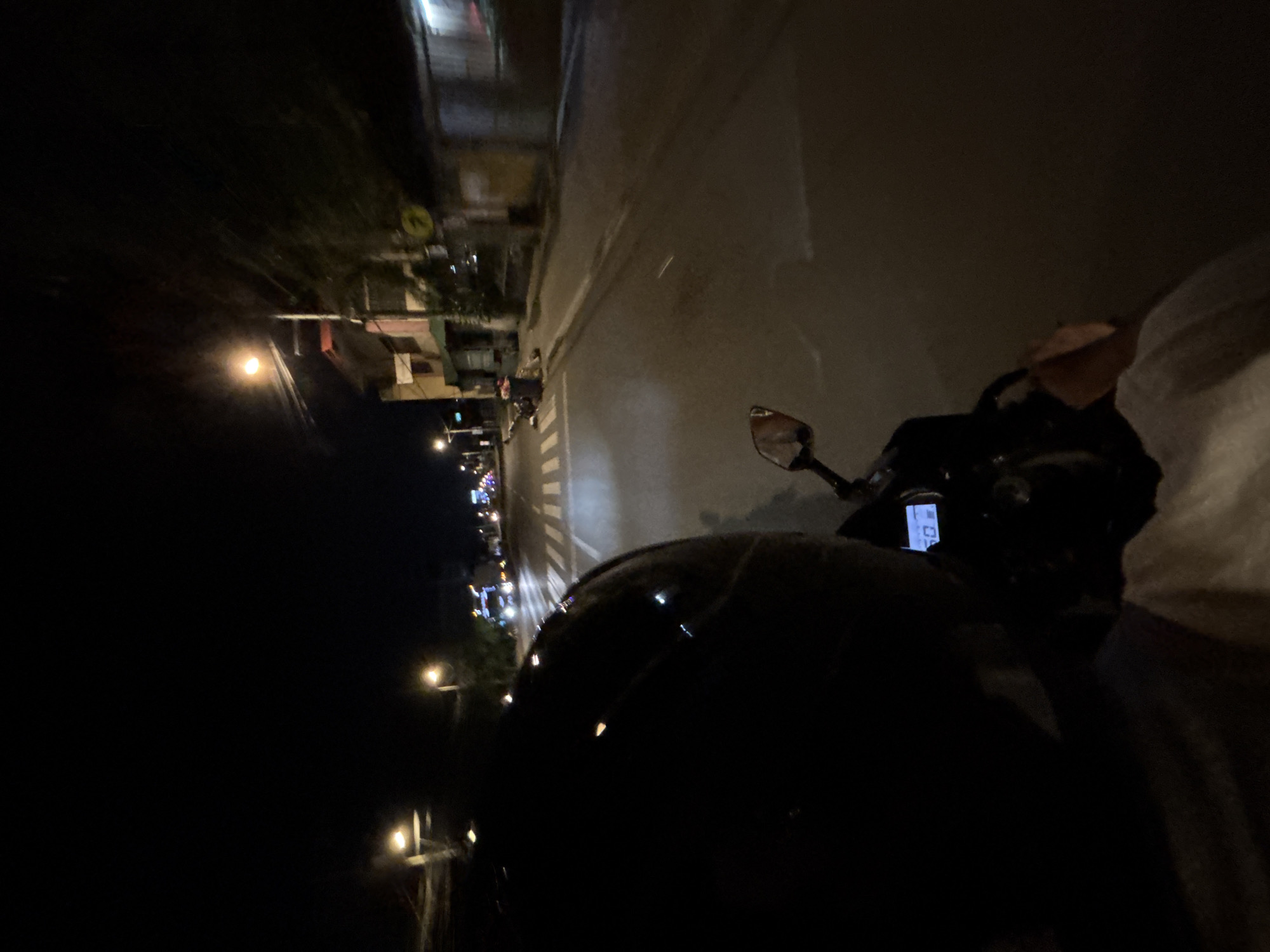 The image depicts a first-person perspective of someone riding a motorcycle at night on a street in Davao City, Philippines. The rider is wearing a dark helmet, the front of which is visible in the foreground, obscuring their face. The motorcycle's dashboard is partially visible, showing a digital display with the number "50" illuminated in blue, likely indicating the speed. The street is paved and appears to be a main thoroughfare, with streetlights illuminating the scene. On the right side of the road, there are buildings with some lights on, suggesting businesses or residences. On the left side of the road, there are more buildings and streetlights. Pedestrian crossing lines are visible on the road. In the distance, headlights of other vehicles can be seen, indicating moderate traffic. The overall environment is urban, characterized by the presence of buildings, streetlights, and vehicles. The time of day is clearly night due to the darkness and artificial lighting.