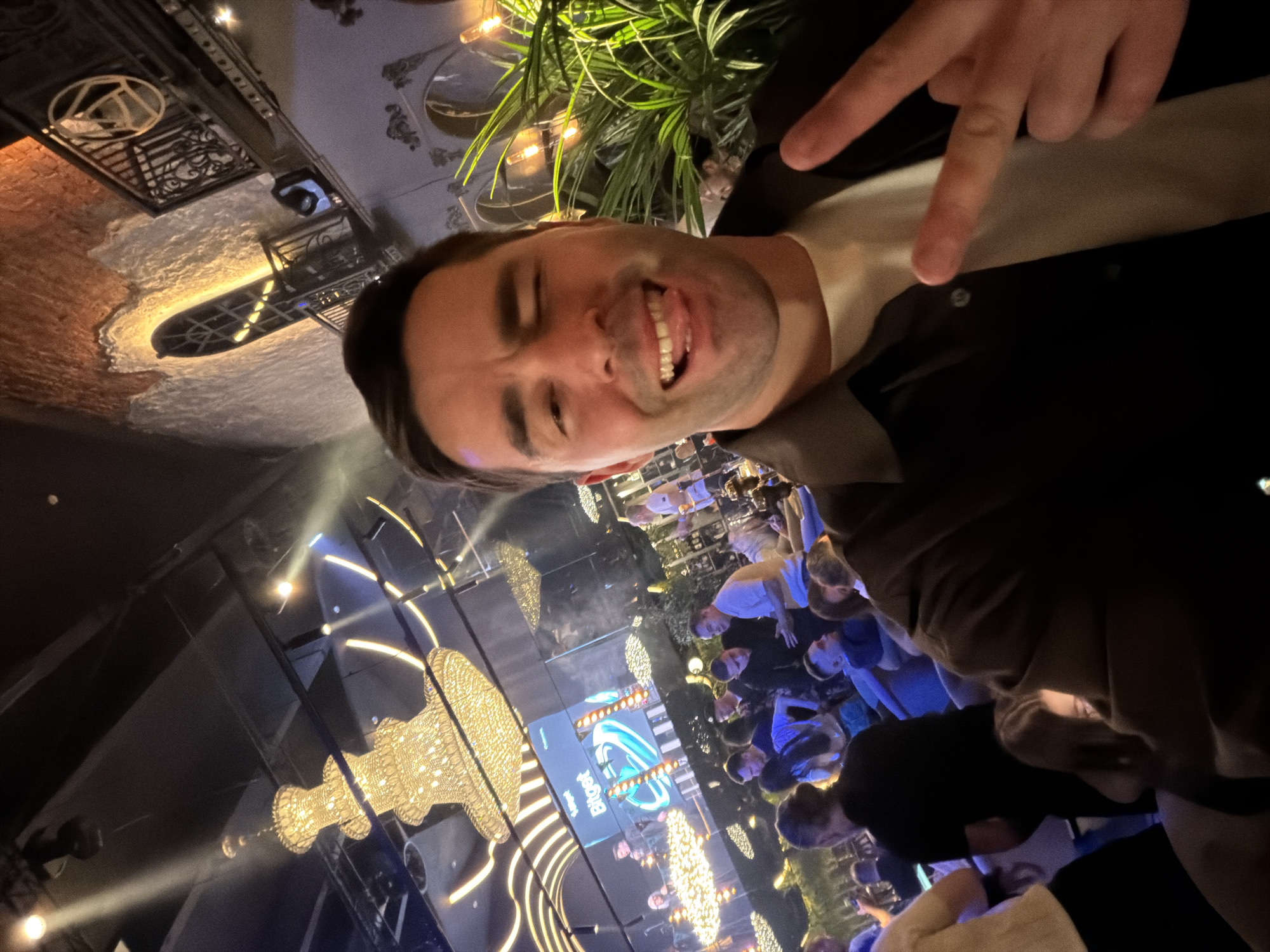 A man takes a selfie in a crowded establishment with a screen displaying the word "Bingo." He is making a peace sign. The location is Moscow, Russia. The establishment appears to be a bar or club with decorative lighting.