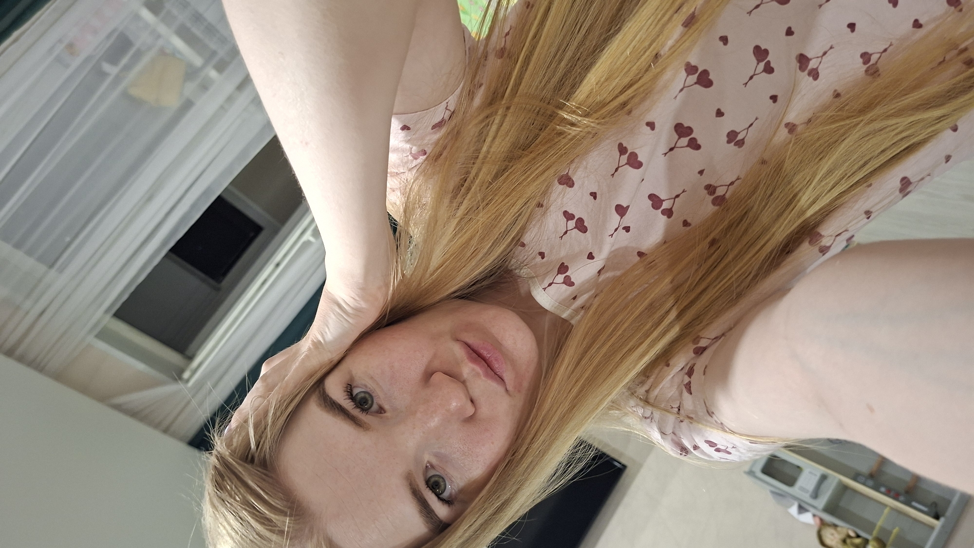 A selfie-style photograph captures a fair-skinned woman with long, strawberry-blonde hair. She is wearing a light pink top with a pattern of small, red hearts. Her hand is resting on her head. The background features a window with a sheer white curtain and what appears to be a skylight. To the right, a partial view of a gray storage unit is visible. The location is Chaikovsky, Russia.