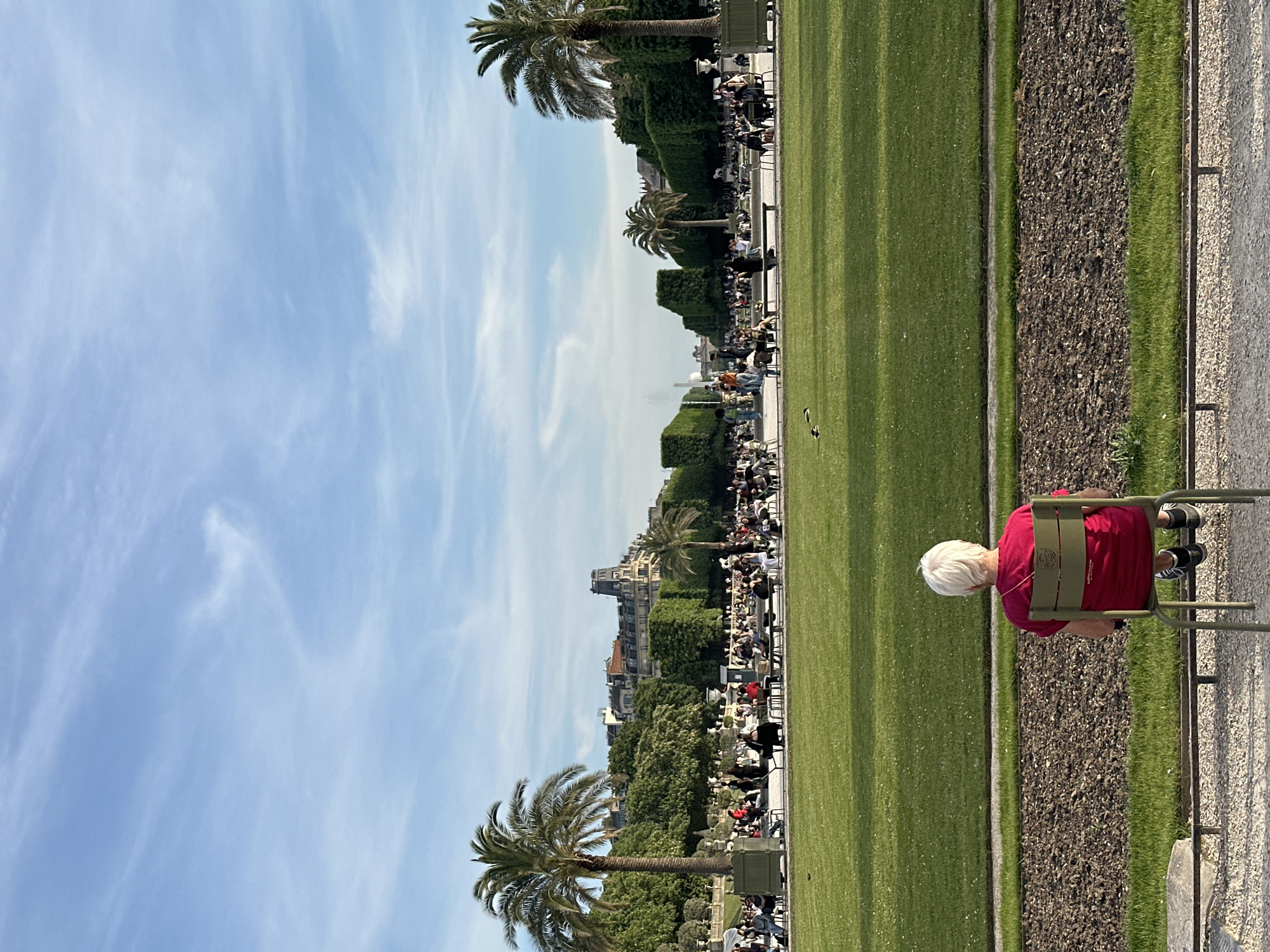 A medium-level shot shows a woman sitting on a bench in the Jardin des Tuileries in Paris, France. She has short white hair and is wearing a red shirt. She is facing away from the camera.
The Jardin des Tuileries is a public garden located between the Louvre Museum and the Place de la Concorde. The garden is filled with green lawns, trees, and sculptures. People are sitting on benches and walking around in the background.
The sky is blue with white clouds. Palm trees are visible.
The Eiffel Tower can be seen in the distance.