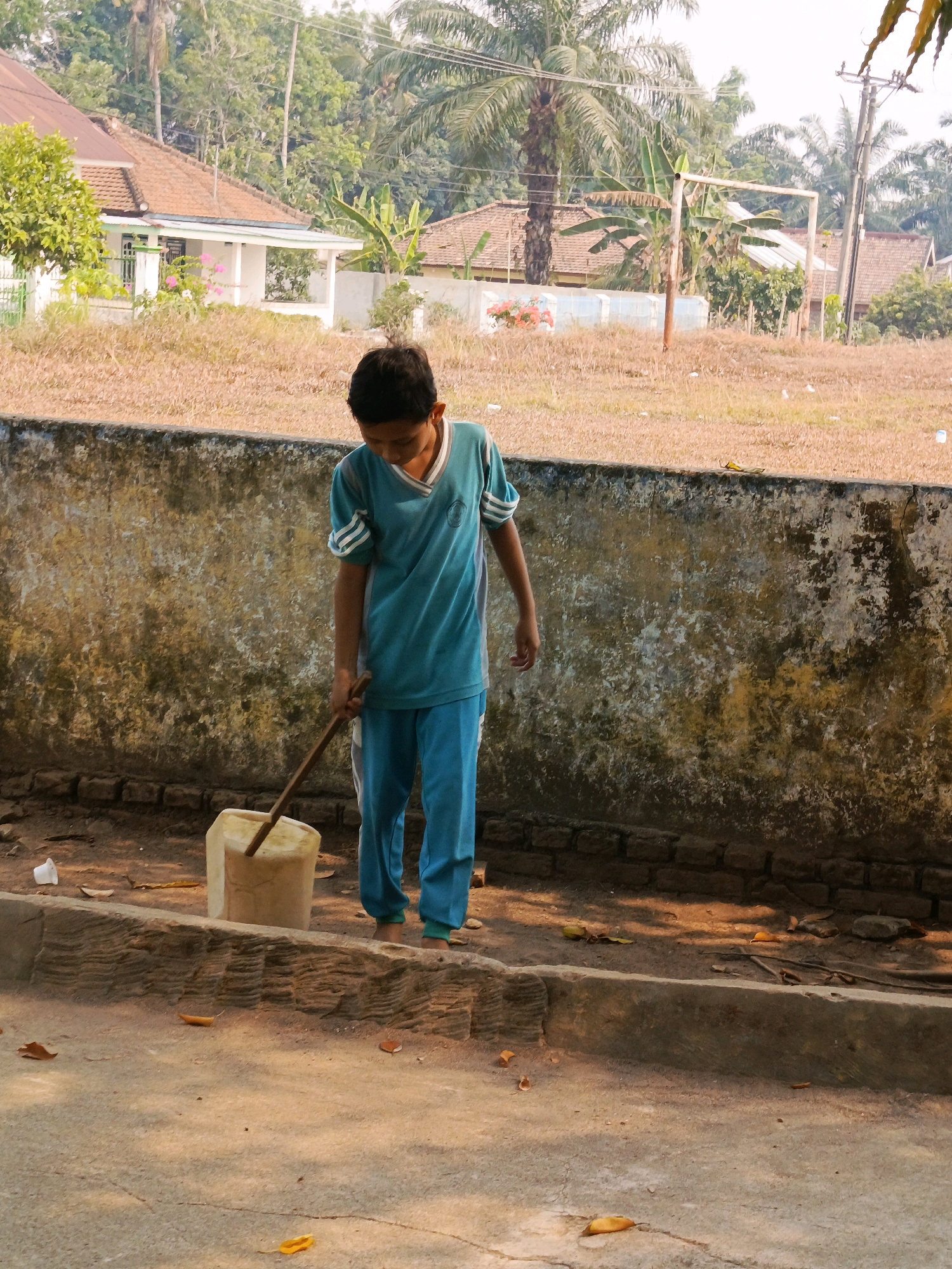 A young boy wearing a teal shirt and blue pants is standing in front of a textured, weathered concrete wall. He is holding a wooden stick, which appears to be attached to a light-colored, cylindrical object. The boy is looking down at the object, and his posture suggests he might be engaged in some task. The ground around him is a mixture of dirt and concrete, with scattered leaves and a white plastic cup visible. In the background, the wall transitions to dry, brownish vegetation. Further back, there are houses with reddish-brown tile roofs and palm trees, indicative of a tropical or subtropical environment. The overall scene suggests a residential or outdoor area, possibly in the city of Karya Dadi, Indonesia, given the presence of tropical flora and architectural styles.