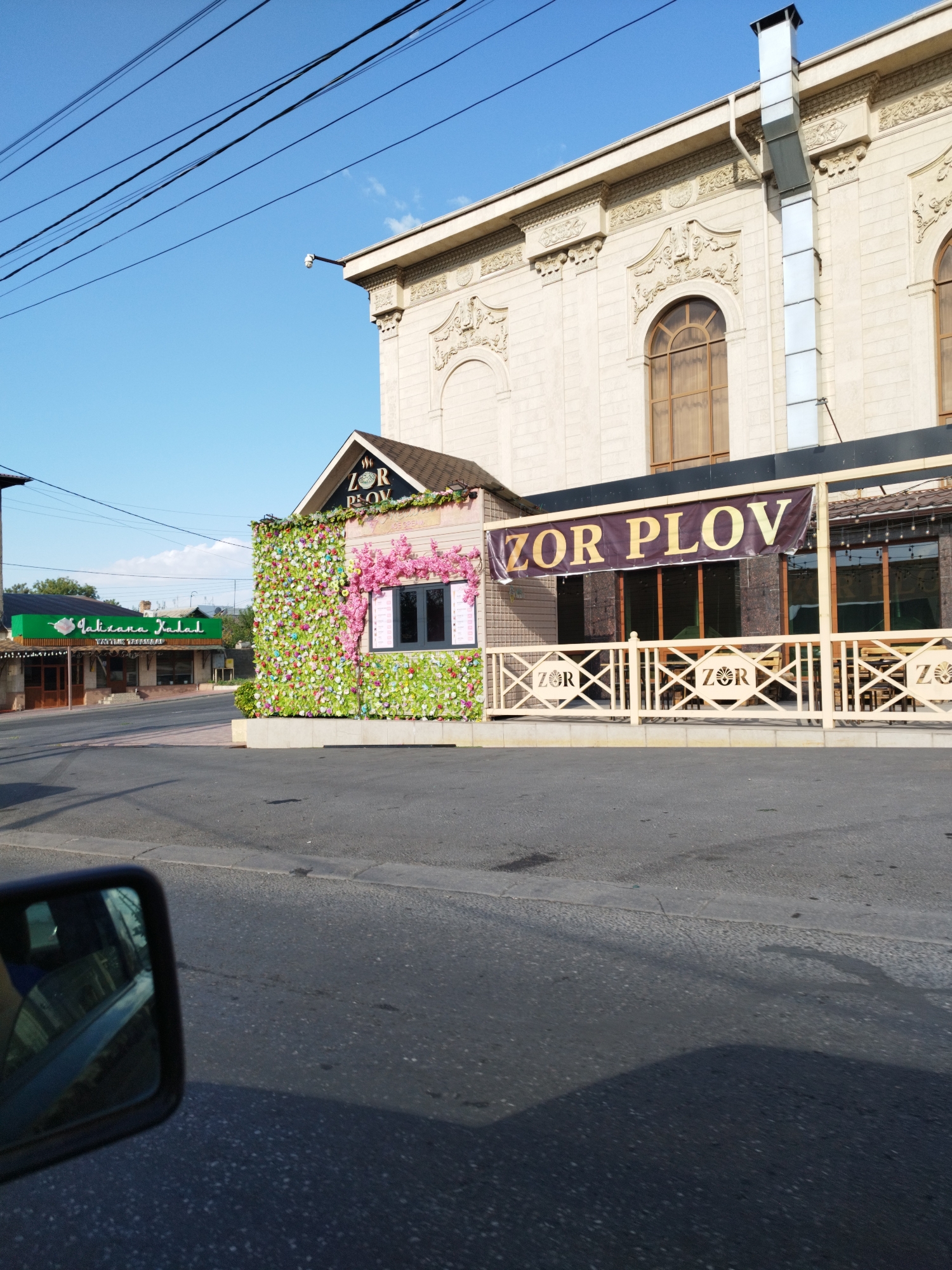 The image captures a street view in Shymkent, Kazakhstan, featuring a restaurant with a distinctive facade. The restaurant, identified by a banner as "ZOR PLOV," is adorned with a vibrant green wall covered in artificial flowers and pink floral accents around a window. Adjacent to it, another establishment with a green awning displays the text "Jalizana Xalal," also appearing to be a food establishment. In the background, a larger building with classical architectural elements, including arched windows and ornate decorations, stands. Power lines are visible overhead against a clear blue sky. The foreground shows a paved road and a car's side mirror reflecting the scene, indicating the photo was taken from a vehicle. No people are visible in the frame.