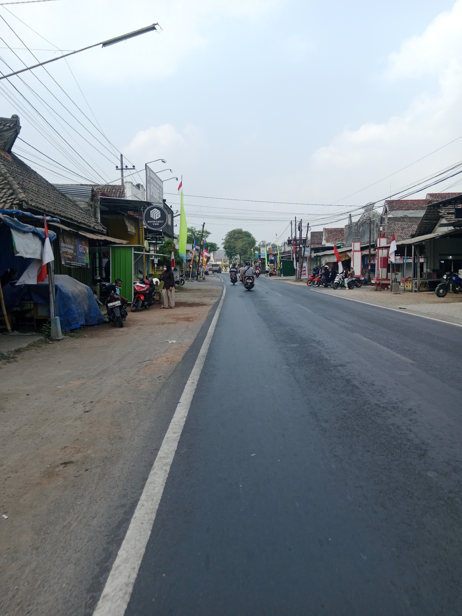The image depicts a street scene in Ringinrejo, Indonesia, under a hazy sky. The road, paved with asphalt, runs towards the horizon, bordered by a dirt shoulder on the left and a line of shops and houses on the right. Several motorcycles are parked on the side of the road and a few are being ridden on the street. Red and white flags, likely the Indonesian national flag, are displayed on some of the buildings. Power lines crisscross above the street, supported by utility poles. The buildings lining the street appear to be single-story structures with tiled roofs, some featuring awnings and signage. A few individuals are visible, some standing near the motorcycles and others on them, engaged in daily activities. The overall impression is of a typical, modest commercial and residential street in a town.