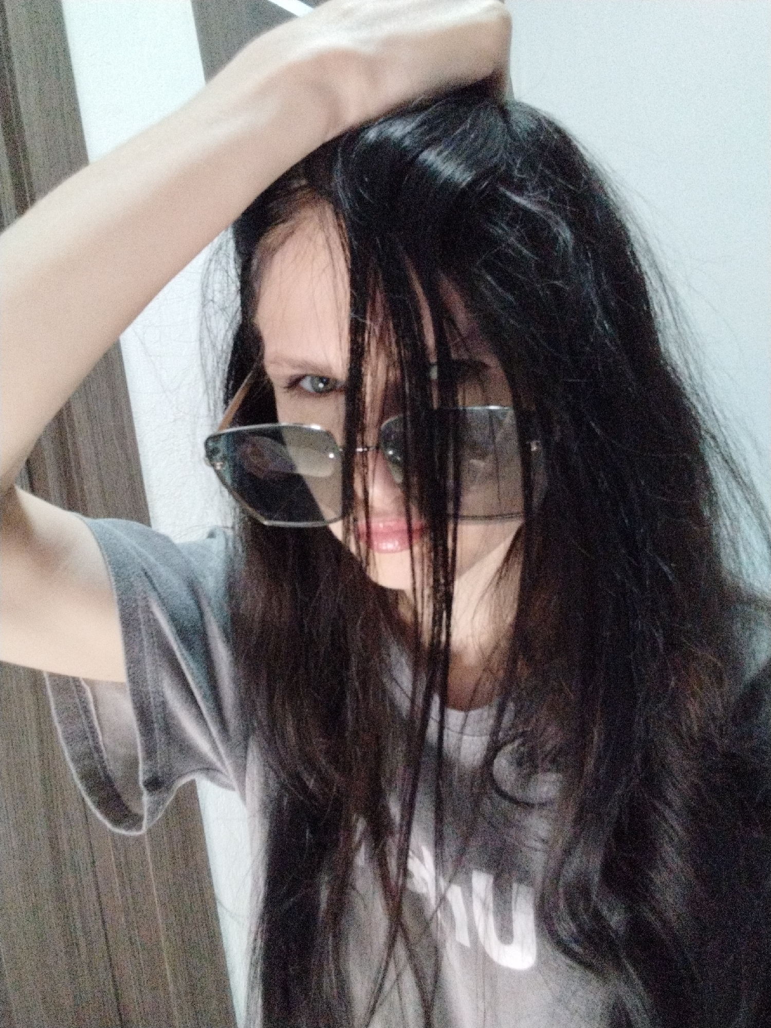 A close-up, waist-up shot shows a young woman with long, dark hair. She is wearing a gray t-shirt and large, angular sunglasses that obscure most of her face. Her right arm is raised, with her hand tangled in her hair. Her left arm is partially visible, resting near her shoulder. The background is softly lit and appears to be an indoor setting, with a vertical wooden panel on the left and a white wall on the right. The lighting suggests it could be daytime, but no specific time can be determined. The overall mood is casual and somewhat mysterious due to the obscured facial features. Some text is visible on the woman's t-shirt, appearing to be the letters "MU" in white block font.