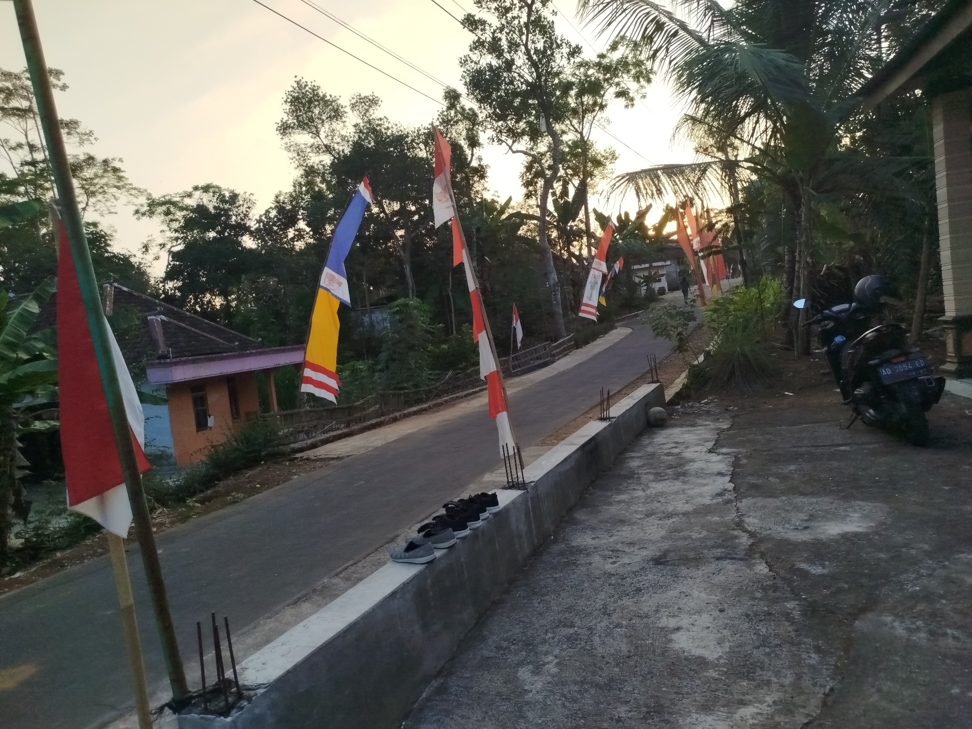 The image depicts a rural street scene in Sewurejo, Indonesia. Several flags, red and white, are displayed on poles along the side of the road, some with blue and yellow accents. The road itself is paved and curves uphill. On the left side of the road, there is a small building with an orange-toned wall and a dark roof. In the foreground, a concrete barrier runs alongside the road, and a row of shoes, including sneakers and sandals, are neatly placed on top of it. Further down the road on the right, a motorcycle with a helmet on the seat is parked. The environment is surrounded by lush greenery, with various trees and vegetation. The sky is a soft, hazy yellow, indicating it might be dawn or dusk.