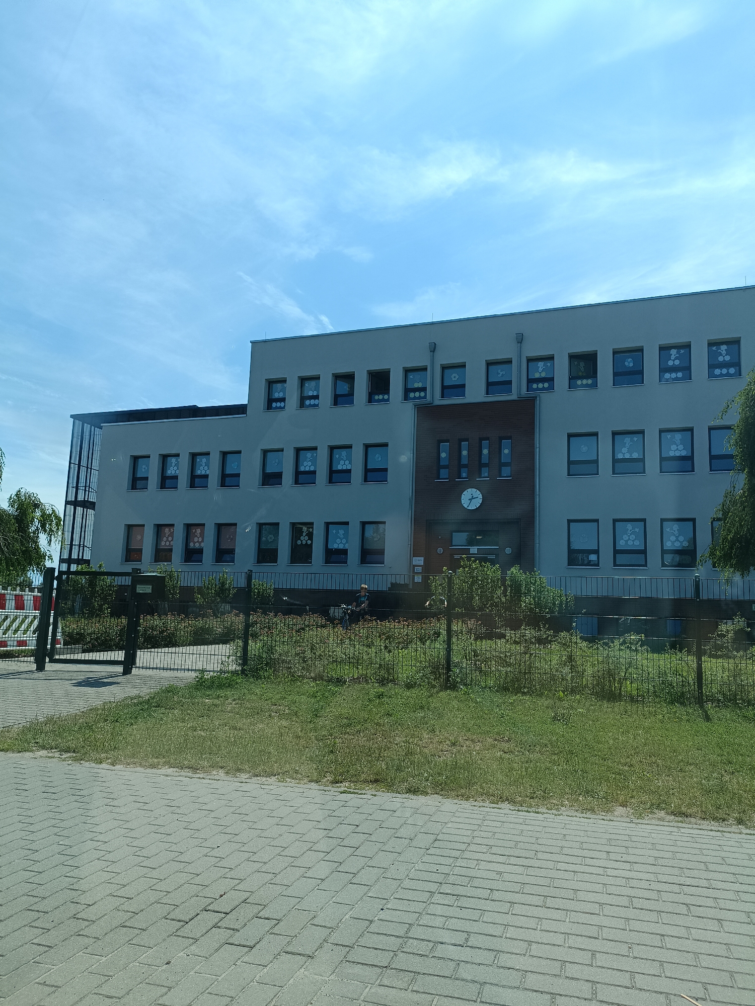 This media file shows a multi-story building, likely a school or administrative building, under a bright, partly cloudy sky. The building has a modern design with a light-colored facade and many rectangular windows. Some of the windows appear to have decorations or displays inside, such as floral patterns or figures. A prominent clock face is visible on the central section of the building, indicating it might be a public building with a clock tower feature.
In the foreground, a paved pathway made of grey interlocking bricks leads towards the building. A metal fence separates the pathway from a grassy area and the building itself. Beyond the fence, there is more greenery and some bushes. On the right side of the image, a person is visible near the fence, possibly interacting with a bicycle. There are also some traffic barriers on the left side of the image, near a gate. The overall scene suggests an outdoor view of a well-maintained public space on a clear day. The time of day appears to be daytime, given the bright lighting and the shadow cast by the building. The weather is fair, with scattered white clouds in a blue sky. The location is identified as Gartz (Oder), Germany. No visible text is present in the image.