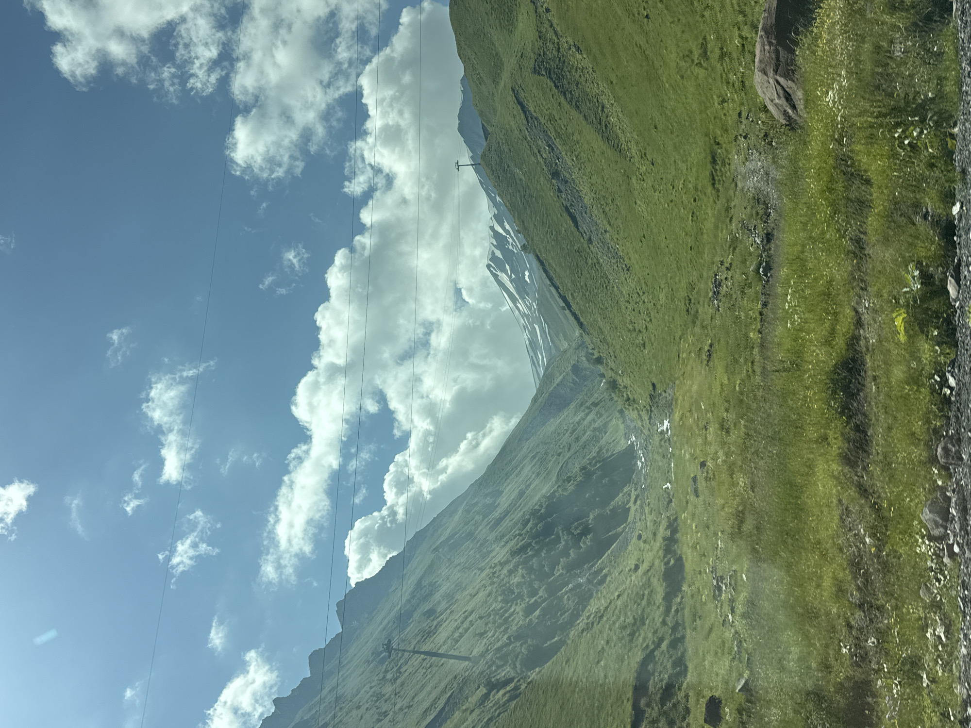 The image shows a lush green valley in the Almasiani region of Georgia. Steep, grassy mountainsides flank a winding river that flows through the valley floor. The sky is bright blue with scattered white clouds, and the sun casts shadows across the landscape. In the distance, snow-capped peaks are visible. There are no people or animals in the image. Electrical wires stretch across the frame, suggesting the presence of human infrastructure in the area. The overall scene is one of natural beauty and tranquility.