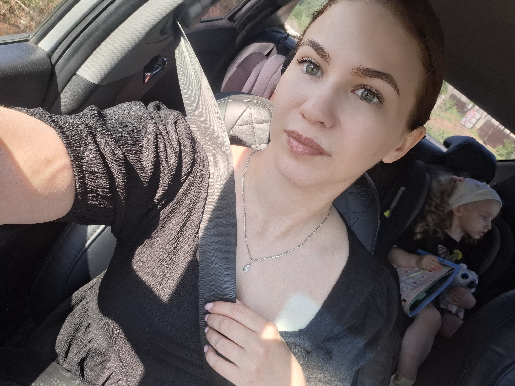 A woman is taking a selfie in a car. She is wearing a black dress, a seatbelt, and a silver necklace. A young child with curly hair and a white head covering is in a car seat in the background, holding a stuffed animal and a book. The car is located near Nizhnyaya Gar', Russia, based on the metadata. The lighting suggests it is daytime.