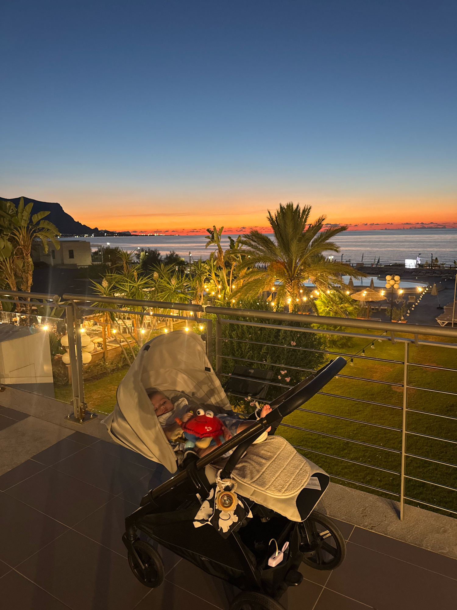 An overhead shot captures a baby in a stroller on a balcony at dusk. The baby is nestled in a light grey stroller cover, propped up with a red crab plush toy. A view of the Mediterranean Sea unfolds in the background, with a sliver of orange and yellow sunset sky visible above the horizon. Palm trees and string lights adorn the scene, illuminating a pathway leading to an outdoor seating area and swimming pool. The location is identified as Isola delle Femmine, Italy.