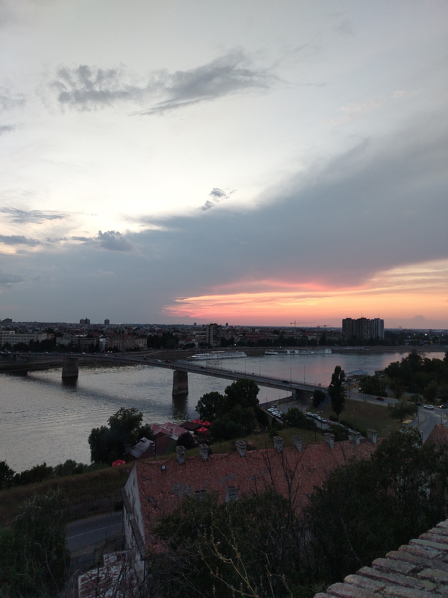 An outdoor, eye-level, medium-shot photograph depicts a city skyline at dusk, with a wide river in the foreground. The sky is a gradient of pale gray clouds at the top, transitioning to hues of pink and orange closer to the horizon. A bridge with multiple concrete supports spans the river, with cars visible on its surface. A large riverboat is positioned on the water to the right of the bridge. The city itself is characterized by a dense collection of buildings, including apartment complexes and smaller structures with red tile roofs, some of which are partially obscured by trees. The foreground shows a grassy embankment leading down to the river, with more vegetation and a brick wall on the far right. The scene is likely Petrovaradin, Serbia, given the architectural style and the presence of the Danube River. There are no identifiable people in the image. The overall atmosphere is serene, bathed in the soft light of sunset.