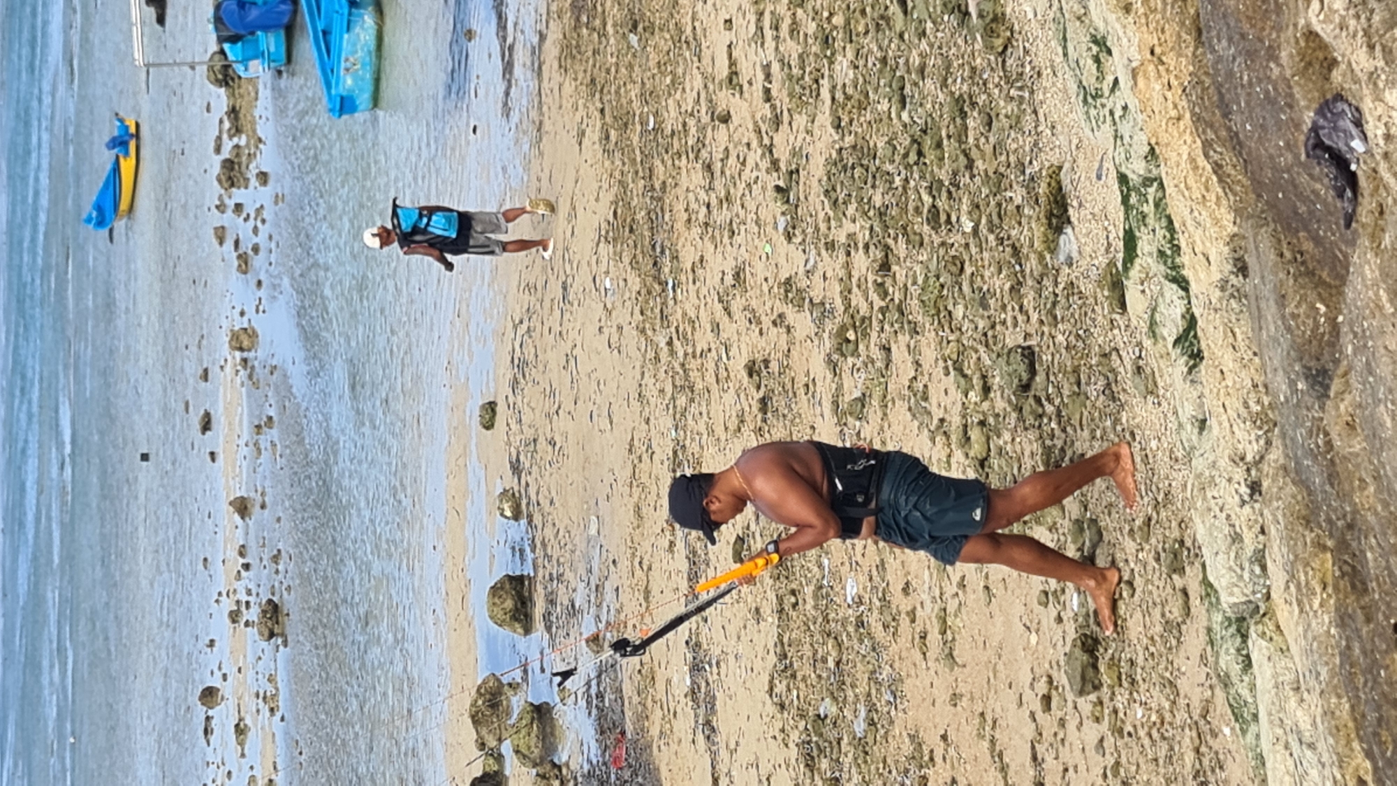 Two men are on a beach in Denpasar, Indonesia. One man, wearing a life vest and a white cap, stands in the shallow water. He is holding a yellow and blue object, possibly a paddleboard or kayak. Another man, wearing a black cap and dark shorts, is on the sand near the water's edge. He is holding a yellow and black kiteboarding bar. The beach is sandy with rocks and some debris scattered around. The water is calm and appears shallow. The time of day seems to be daytime, likely morning or afternoon, with bright sunlight. The weather appears to be clear and pleasant for outdoor activities. The scene suggests a recreational setting, likely for water sports.