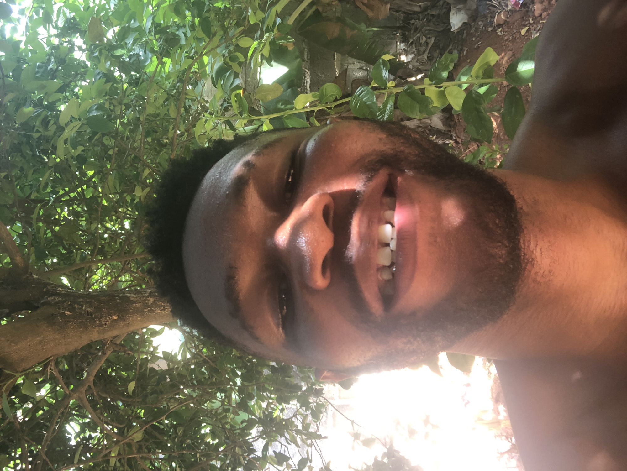 A close-up, tilted upward selfie photograph shows a Black male individual with short, dark hair, facial hair, and visible perspiration on his face. He is smiling, revealing his upper teeth. The background consists of green foliage, possibly trees, with some light breaking through. Some branches are visible, along with what appears to be some sort of structure in the background, possibly a building of some kind.
The location is labeled as "Unknown, Unknown."