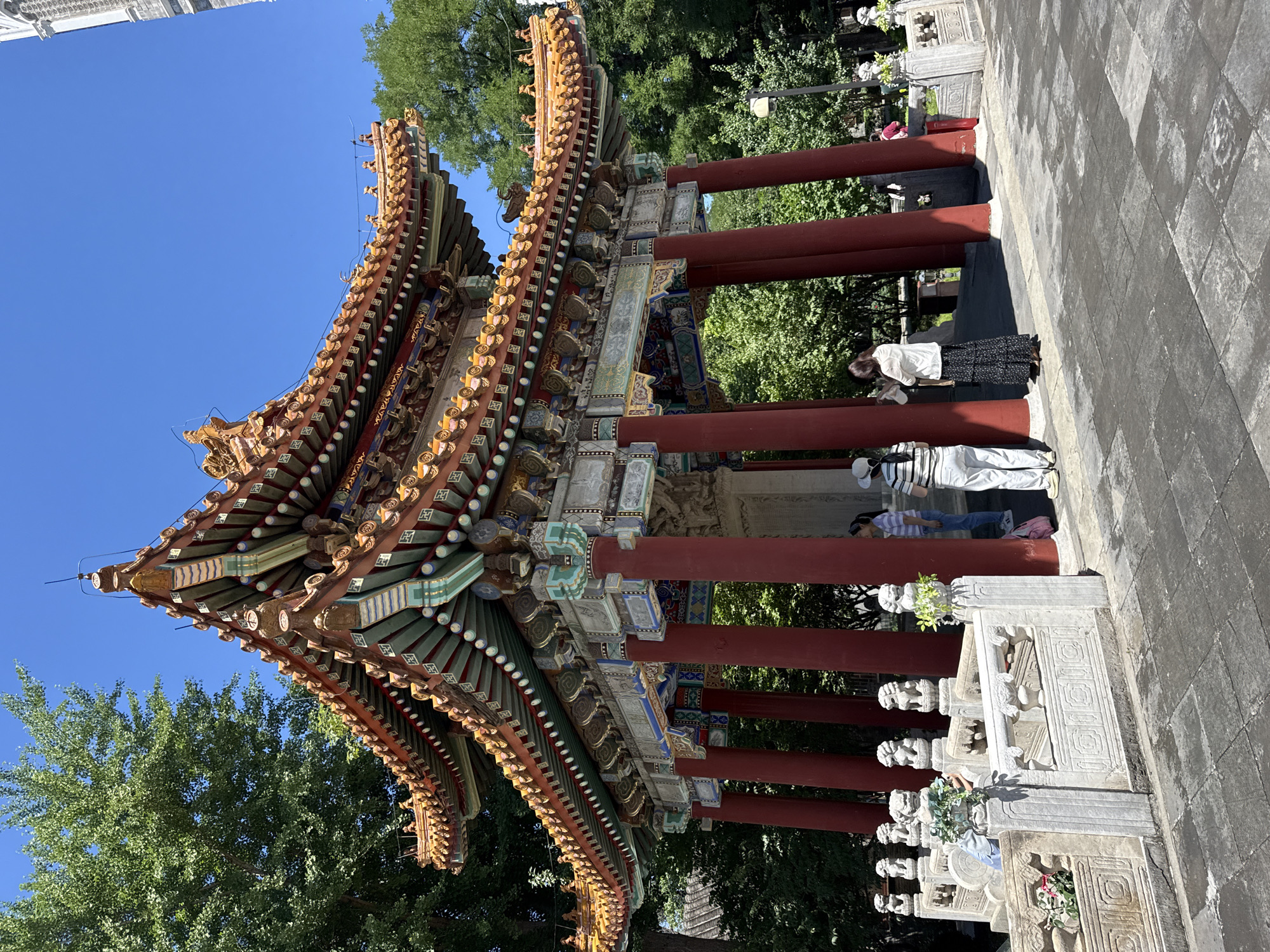 This detailed image captures a vibrant, multi-tiered traditional Chinese pavilion located in Xicheng District, Beijing, China, likely within Beihai Park. The pavilion features eight robust, round, crimson-red support pillars. Above the pillars, an elaborate frieze and beam structure exhibit intricate traditional Chinese artwork, predominantly in blue, green, red, and gold patterns against a white and grey background. The pavilion is crowned with a multi-eaved, hip-and-gable roof covered in bright orange-red glazed tiles, accented with yellow and green glazed elements. The upturned eaves are decorated with numerous small, mythical animal figurines and intricate patterns. The underside of the eaves reveals complex Dougong (bracket sets) painted in greens, blues, reds, and gold, indicative of imperial architectural style.
Inside the pavilion, a large, rectangular stone stele is visible, partially obscured by visitors and shadows, suggesting it rests on a traditional tortoise-shaped base.
The pavilion stands on a paved area composed of large, dark grey, rectangular stone slabs showing signs of wear. To the left and partially extending in front of the pavilion is a white stone balustrade. This balustrade features carved panels and posts, each topped with a small, stylized stone lion sculpture. Small arrangements of white and green artificial flowers are placed on some of the balustrade posts.
The pavilion is surrounded by lush, dense green trees, suggesting a park-like setting, under a clear, bright blue sky. The scene is illuminated by strong natural sunlight, casting distinct shadows from the pavilion's structure and the people present.
Approximately eight individuals are visible around and within the pavilion. In the foreground, three women are prominent: one woman, viewed from behind, wears a white and black striped shirt, white pants, and a white baseball cap; another woman wears a long-sleeved white top and a long black skirt with a floral pattern, observing the stele; a third woman, also viewed from behind, wears a light blue striped top and blue jeans, standing near a pillar. Other individuals are partially visible further back, appearing to be tourists or visitors observing the structure. Their attire is casual. A dark street lamp post is partially visible amidst the trees in the far right background.
