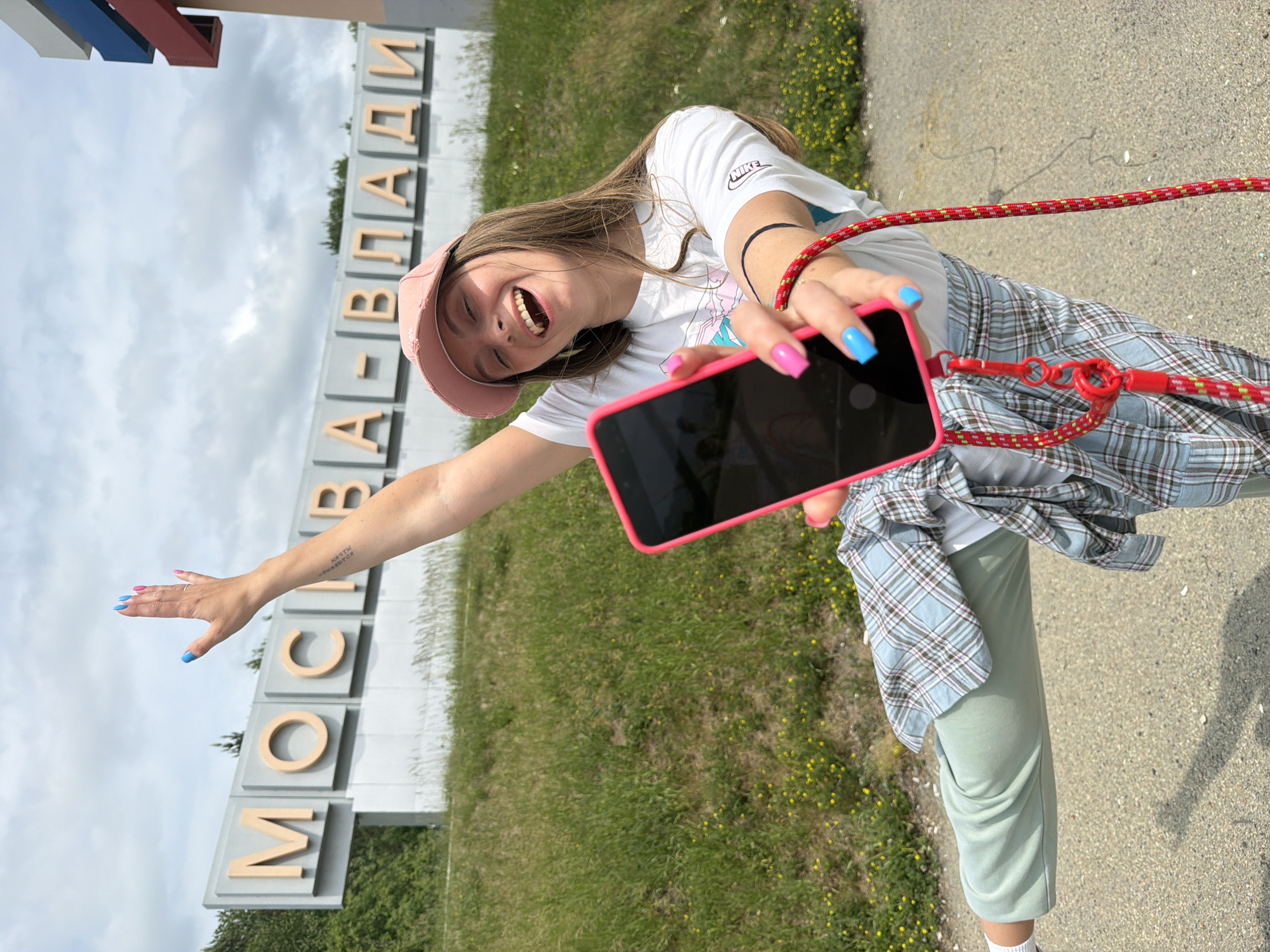 The image shows a young woman with long brown hair, wearing a pink baseball cap, a white t-shirt, a plaid shirt tied around her waist, and light green sweatpants. She is holding a pink-cased smartphone in her right hand, which is outstretched towards the camera. Her left arm is raised, with her hand waving. She has bright pink and blue fingernail polish. The background features a large sign with Cyrillic lettering that reads "МОСКВА-ВЛАДИ", indicating the location is likely near Moscow, Russia. The sign is set against a backdrop of a cloudy sky and a grassy slope with some scattered yellow flowers. The woman is positioned on a paved surface, possibly a road or pathway, with a grassy embankment behind her.