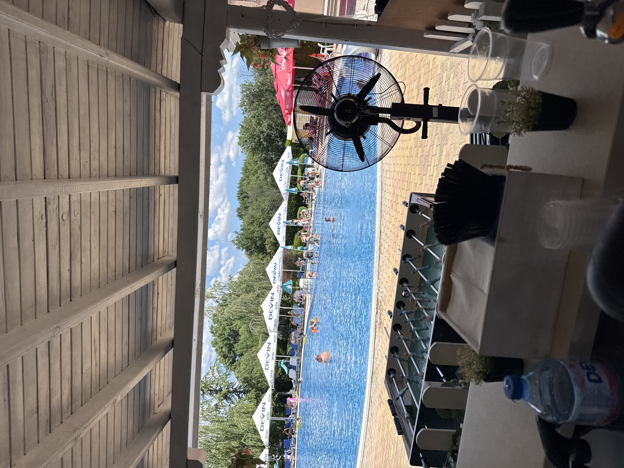 The image depicts an outdoor scene at a swimming pool, likely a resort or water park, in Yagoda, Bulgaria. The foreground features a foosball table and a bar area with a plastic water bottle and cups. A large black industrial fan is positioned in front of the pool, suggesting a warm day.
The mid-ground reveals a bright blue swimming pool filled with people enjoying the water. Several individuals are swimming, and some appear to be relaxing on inflatable devices. Around the pool, rows of lounge chairs are visible, some occupied by sunbathers. Above the lounge chairs, white tents with the "DEVIN" logo provide shade.
The background consists of lush green trees and a blue sky with scattered white clouds, indicating pleasant weather. The overall atmosphere suggests a leisurely day at a recreational facility. The architecture visible, such as the wooden roof structure, hints at a covered outdoor area designed for patrons to relax in.