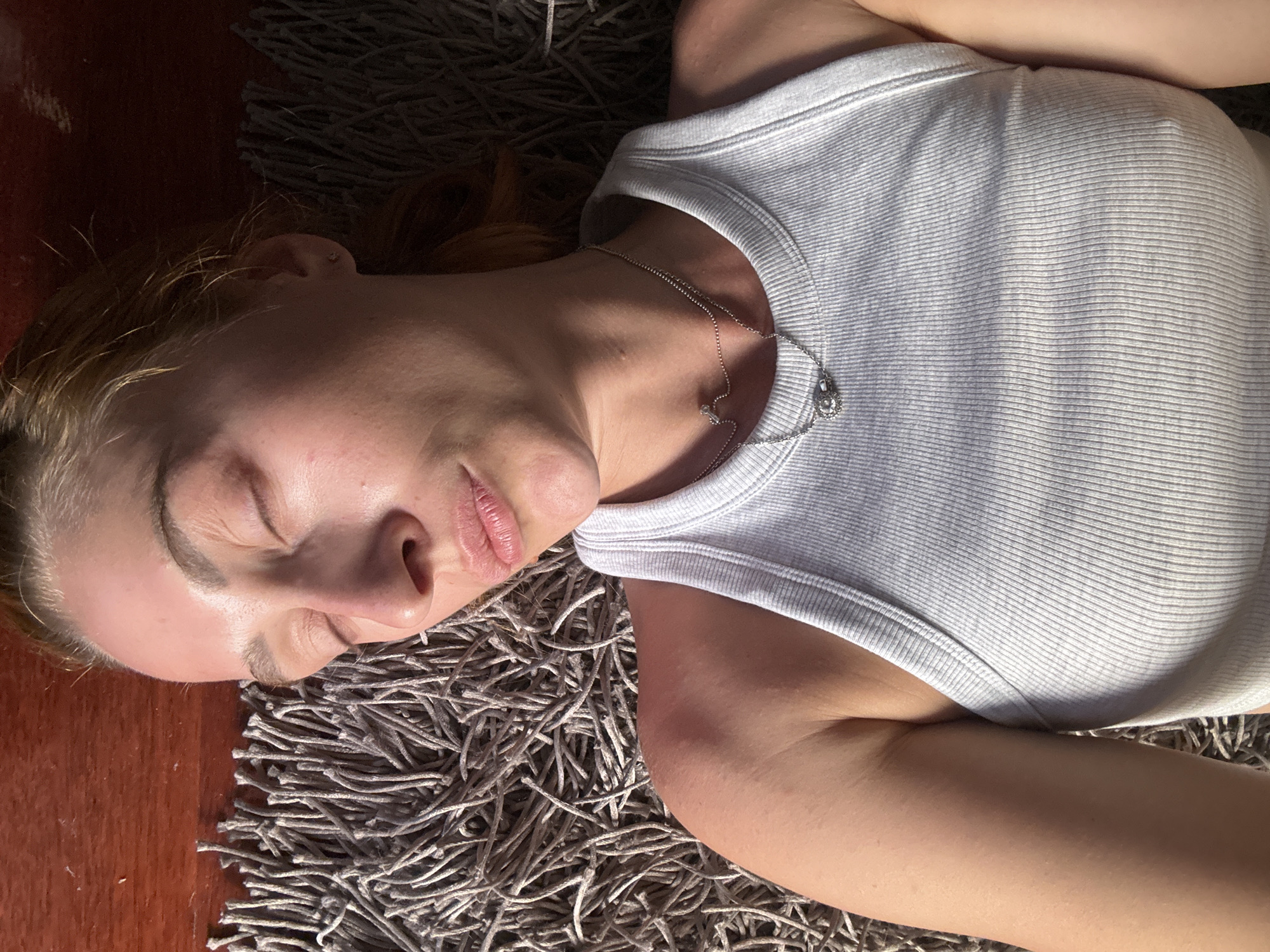 A fair-skinned individual, likely female, with light brown hair, is depicted lying supine with eyes closed, suggesting rest or sleep. They are wearing a light grey, ribbed tank top and a layered silver necklace featuring two distinct pendants, one smaller and round, the other a larger, intricate design. A small, singular stud earring is visible in the right ear. The skin on the exposed arm and shoulder exhibits a tanned appearance. The individual is positioned on a shaggy, grey textured rug, with a section of smooth, reddish-brown wooden flooring visible in the upper-left quadrant of the frame. The scene is illuminated by strong, directional light, creating distinct highlights and shadows, consistent with natural sunlight. The location is Budva, Montenegro.