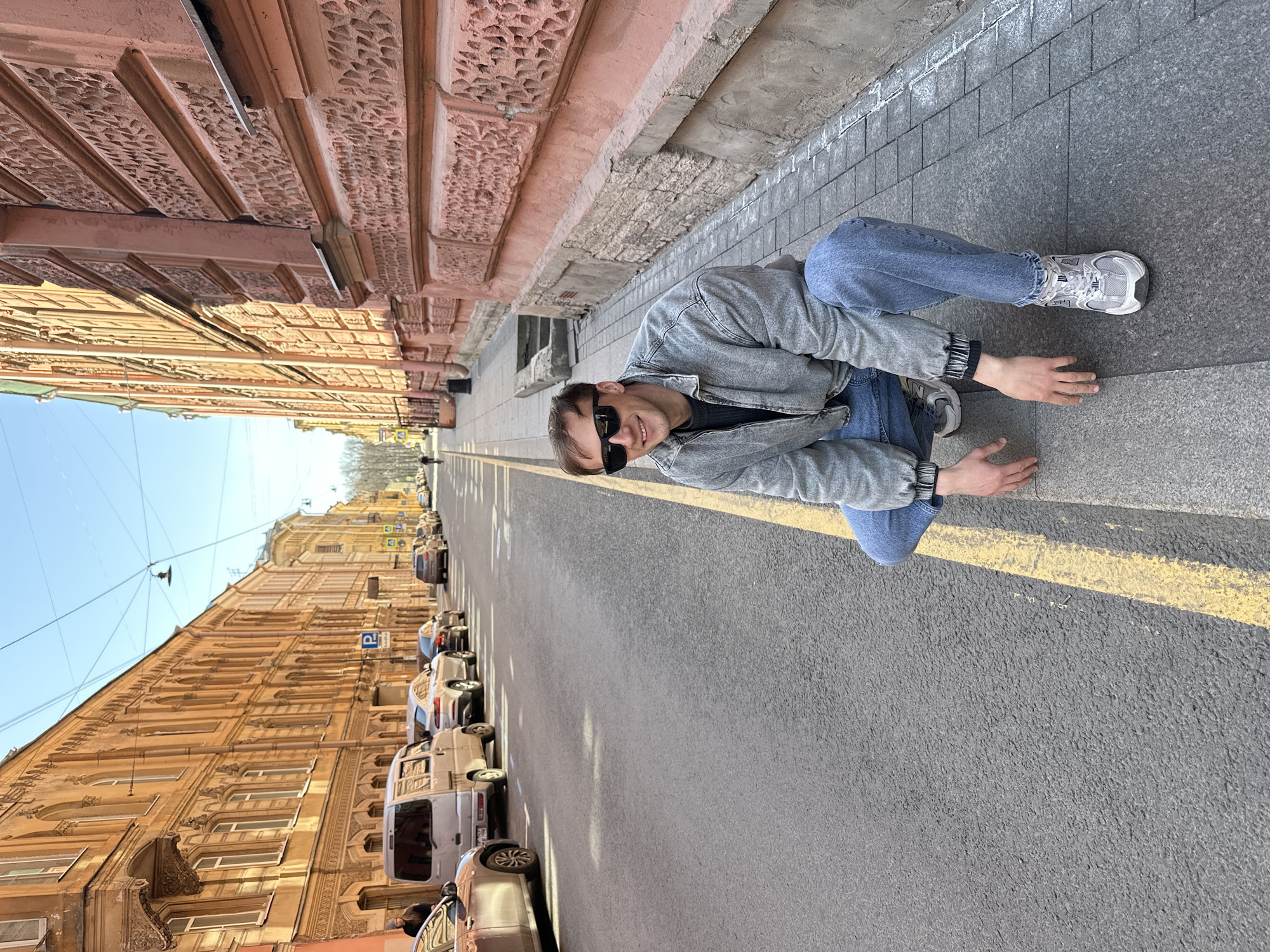 The image shows a young man squatting on a sidewalk in an urban setting, identified as Saint Petersburg, Russia. He is positioned on the right side of the frame, smiling and looking towards the camera. He wears black sunglasses, a grey denim jacket over a dark long-sleeved top, blue jeans, and grey and white New Balance athletic shoes.
The scene depicts a street bathed in bright sunlight under a clear blue sky. On the right, behind the man, stands a multi-story building with a reddish-brown, highly ornate facade, featuring intricate carved patterns and decorative architectural details. On the opposite side of the street, a long, multi-story building with a yellowish-brown facade also displays classical architectural elements, numerous windows, and cornices.
The man is on a grey paving stone sidewalk, adjacent to an asphalt road. A bright yellow line is painted along the edge of the road, separating it from the sidewalk. Several cars are parked parallel to the curb on the left side of the street. A blue parking sign with a white "P" is visible further down the street on the left. The activity captured is the man posing for a photograph, suggesting a relaxed and positive mood.