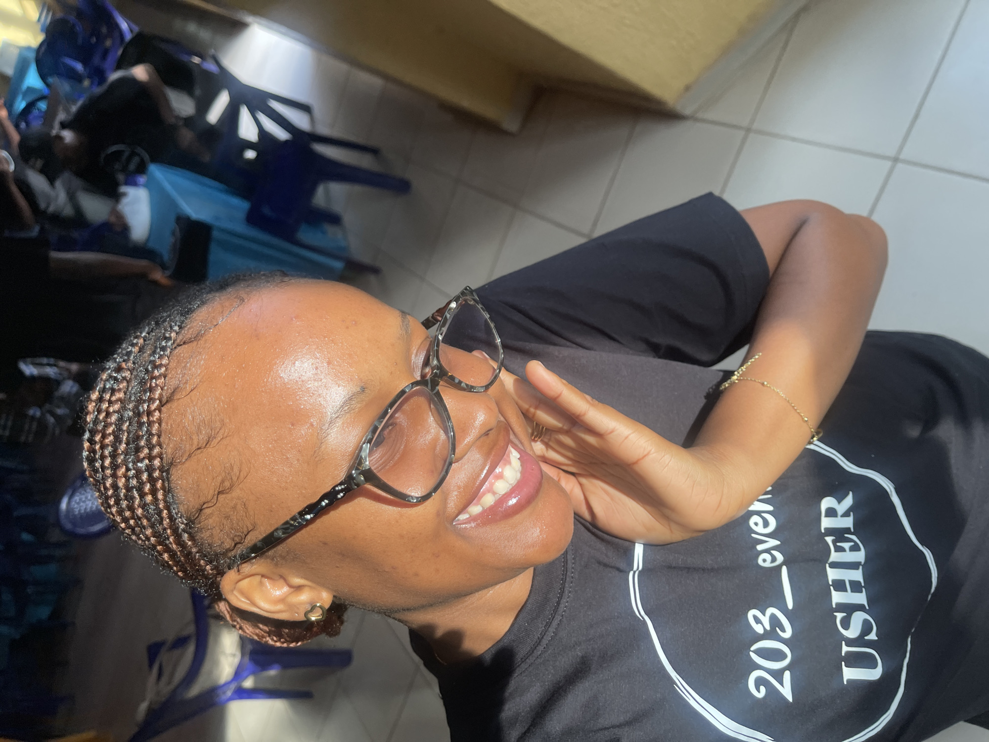 The image shows a close-up, high-angle shot of a young Black woman in a dark t-shirt with "203_even USHER" printed on it in white lettering within a circular design. She is wearing clear-rimmed glasses with a patterned design on the arms and a small gold heart earring. Her right hand is raised to her cheek, and she has a bright smile, with her teeth visible. She is wearing a delicate gold bracelet on her right wrist. The woman's braided hair is visible. The background is out of focus but shows blue chairs, tables draped in a light blue cloth, and a tiled floor. The lighting suggests an indoor setting with natural light entering from the right. The scene appears to be an event or gathering in Bukuru, Nigeria.