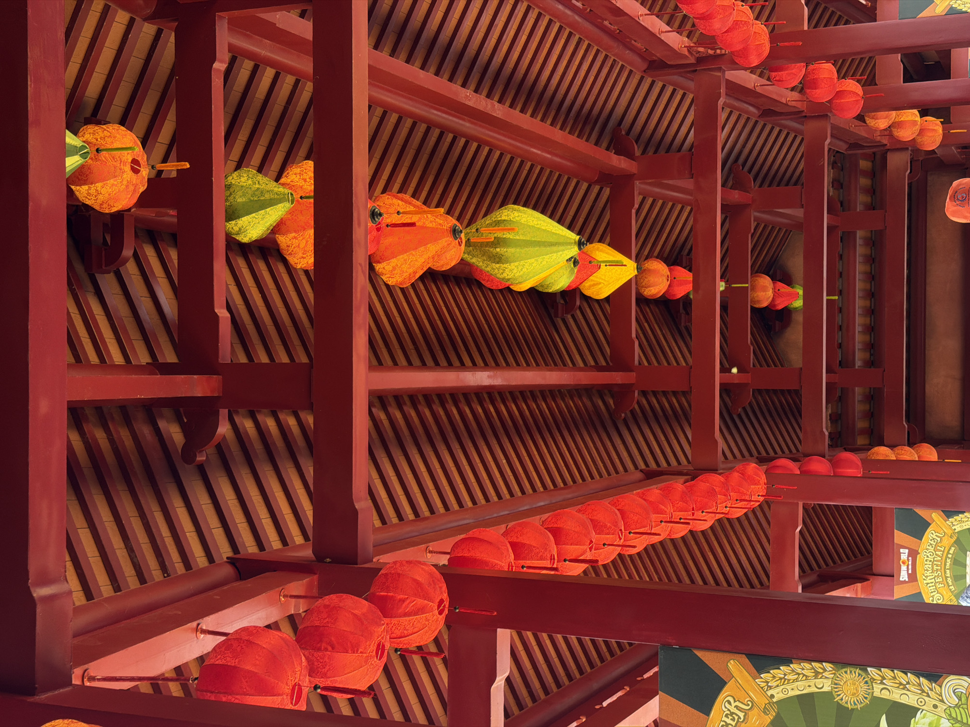 The photograph depicts a view looking upwards at the traditional wooden architecture of a building in Đà Nẵng, Vietnam. The architectural structure is painted red and features a tightly packed pattern of wooden slats on the ceiling. Strings of lanterns hang from the structure. Some lanterns are round and red, while others are more elongated with various hues of orange, yellow, and green. The image is composed with a unique perspective, focusing on the repetitive patterns and vibrant colors of the hanging lanterns against the wooden backdrop. Part of a banner for a beer festival is visible in the lower right corner.