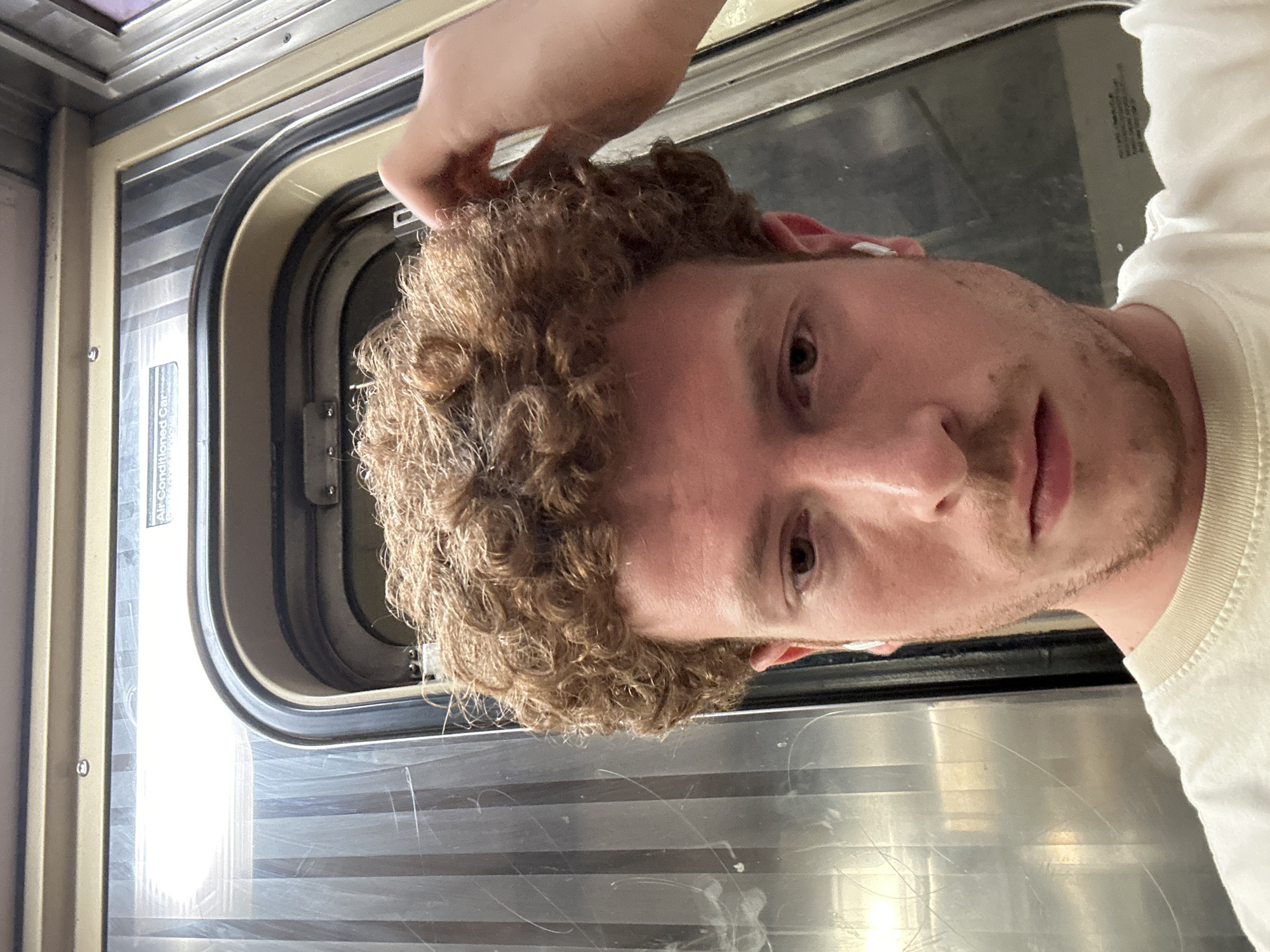A young man is captured in a selfie taken inside what appears to be a train or subway car. He is positioned with his head slightly tilted, looking directly at the camera with a neutral expression. He has light brown, curly hair and some facial stubble around his chin and upper lip. A white earbud is visible in his right ear. His left hand is raised, partially visible near the top of his head, possibly adjusting his hair or holding the camera device. He is wearing a light-colored, likely white or off-white, long-sleeved t-shirt.
The setting behind him is the interior of a vehicle, characterized by a rounded rectangular window or door frame with light-colored (beige/cream) paneling and metallic trim. Through the window, several horizontal, reflective or metallic strips are visible, suggesting closed blinds or a patterned exterior, and a reflection of light is present. On the window frame, a sticker clearly reads "Air Conditioned Car," indicating the type of vehicle. Below this, smaller, indistinct text is also present. The overall environment suggests an older model of public transport vehicle, consistent with a train or subway car in an urban setting like New York, United States. The time of day is not clearly discernible, but interior lighting is present.