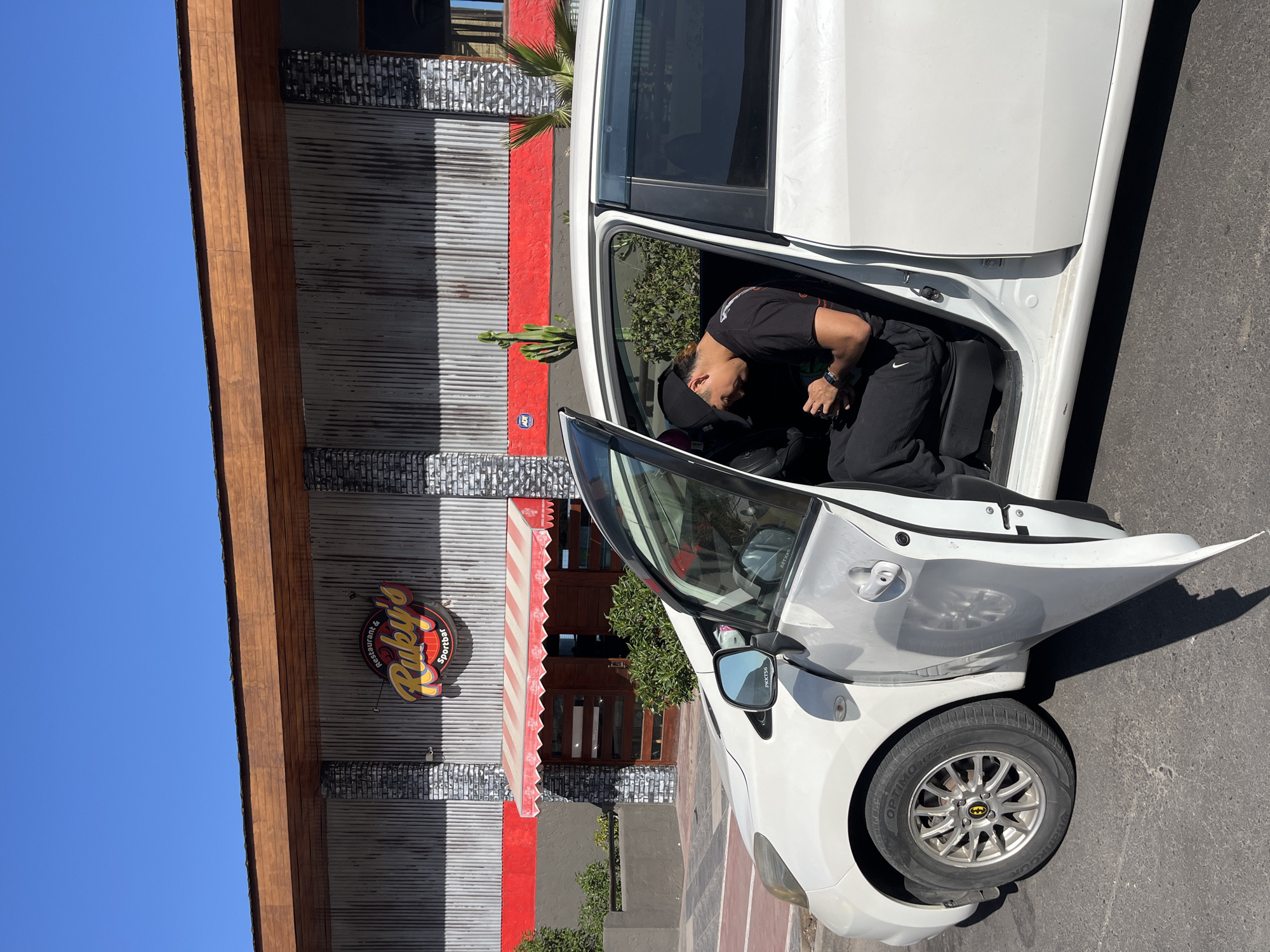 This image, taken on a bright, sunny day in what appears to be Arica, Chile, shows a person interacting with a white car parked outside a commercial building. The scene is illuminated by strong sunlight, casting distinct shadows.
In the foreground, a white 5-door hatchback car, possibly a Toyota Yaris or similar model, is parked on a paved surface. The driver's side door is wide open. A person, appearing to be a man, is leaning into the car's interior. He is dressed casually in a black t-shirt, black tracksuit bottoms with a white Nike swoosh logo visible on the thigh, and a black baseball cap worn backward. A watch is visible on his left wrist. His posture suggests he is either getting into or out of the vehicle, or reaching for something inside. The car's silver multi-spoke wheels feature distinctive black and yellow Batman logos on the center caps. The tires are Hankook Optimo H724. The driver's side mirror has "PUNTEO" written on it.
Behind the car stands a building with a textured facade. It combines horizontal corrugated metal panels with sections painted in a vibrant red and darker, possibly brick-patterned, elements. A robust wooden beam forms part of the roof's eave. A circular sign prominently displayed on the building reads "Ruby's Restaurant & Sportbar" in yellow and red lettering. Below the sign, a red and white striped awning extends over what appears to be a main entrance with wooden double doors. Greenery, including a tall, slender cactus, is visible near the building's base. The sky above is a clear, deep blue.