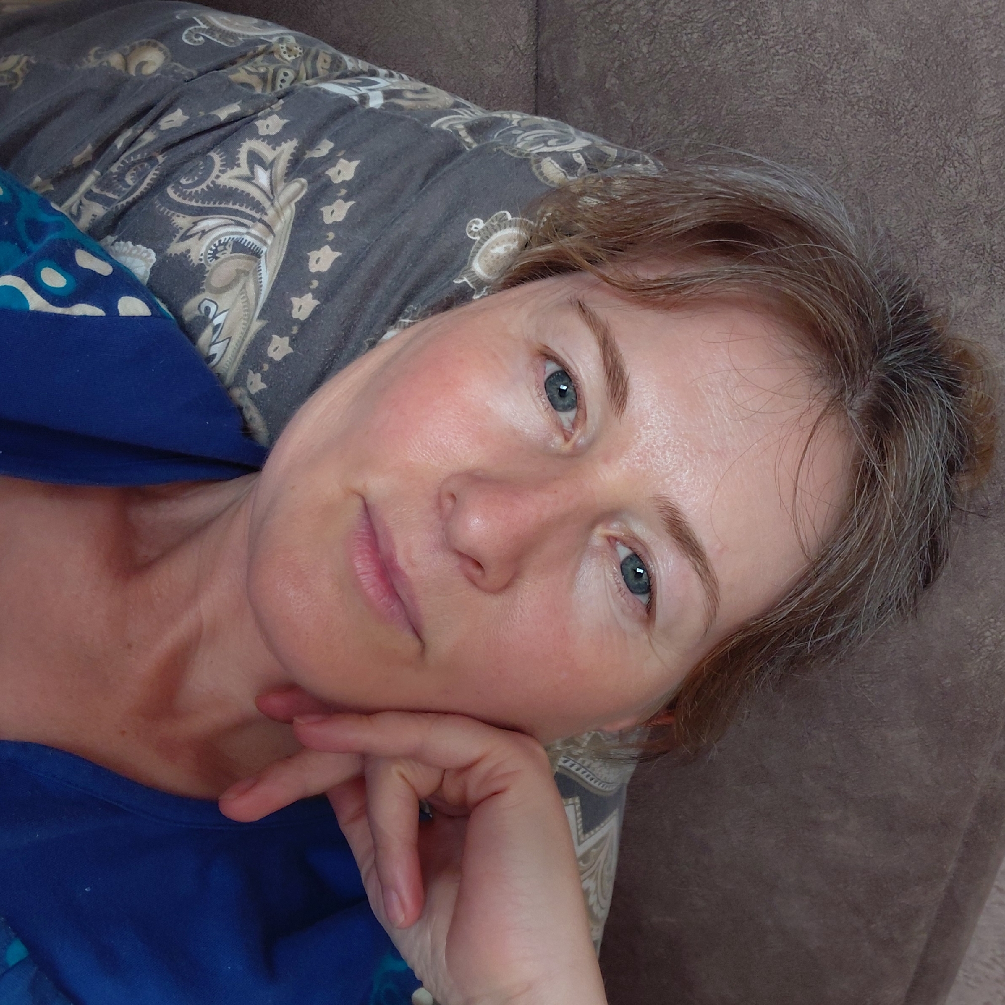 The image is a close-up selfie of a woman lying down and resting her head on a pillow. She has fair skin, light brown hair with some gray strands pulled up in a bun, and blue eyes. Her hand is resting on her face, near her cheek. She is wearing a blue top with a patterned scarf or shawl around her shoulders. The background appears to be a textured brown surface, possibly a couch or sofa. The setting is likely indoors. Without further information, the specific location within Novosibirsk, Russia, cannot be determined.