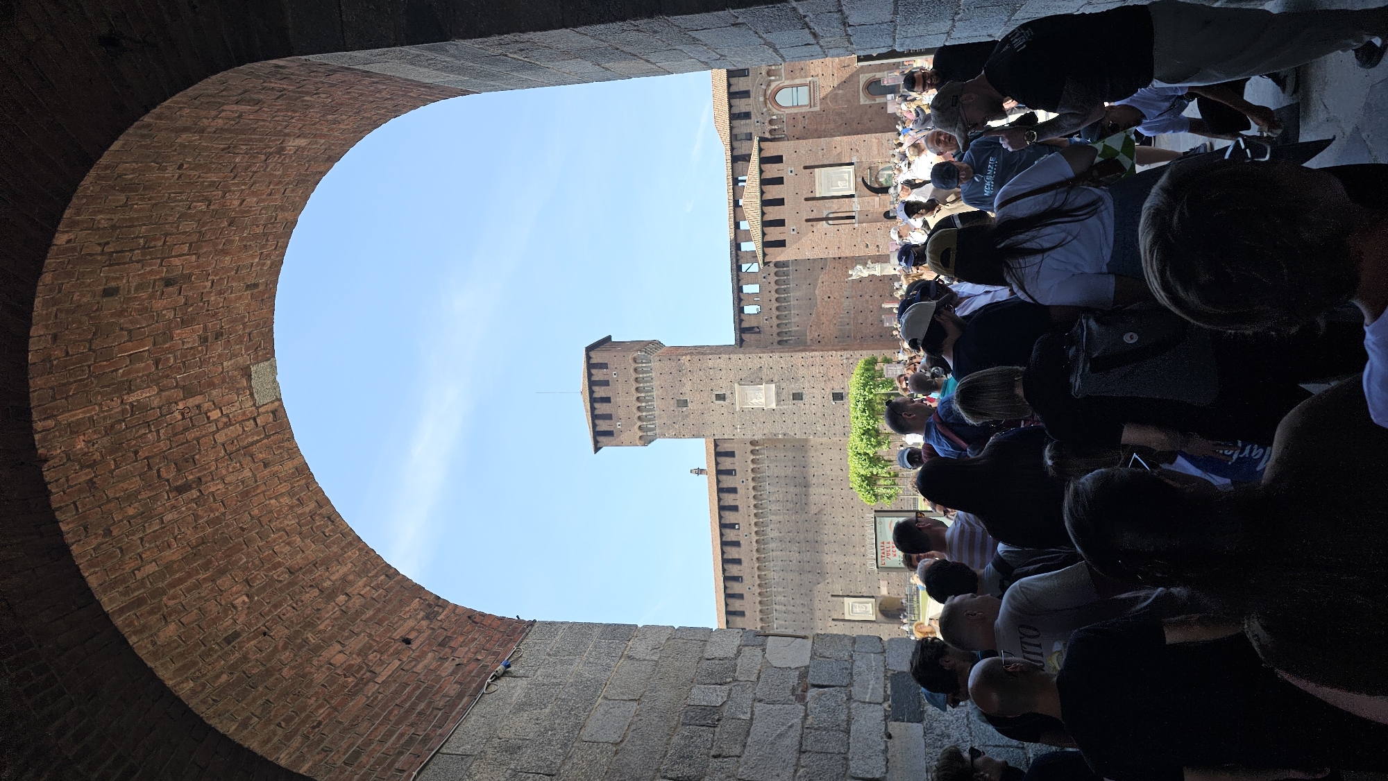 The image captures a partial view of the Sforza Castle in Milan, Italy, as seen through a brick archway. The castle's brick facade and turrets are visible against a clear, blue sky. In front of the castle, a crowd of people is congregated. The foreground is dominated by the stonework of the archway, both the brick of the arch itself and the stone blocks on the side. The image appears to be taken from a low angle, looking upwards and outwards toward the castle.